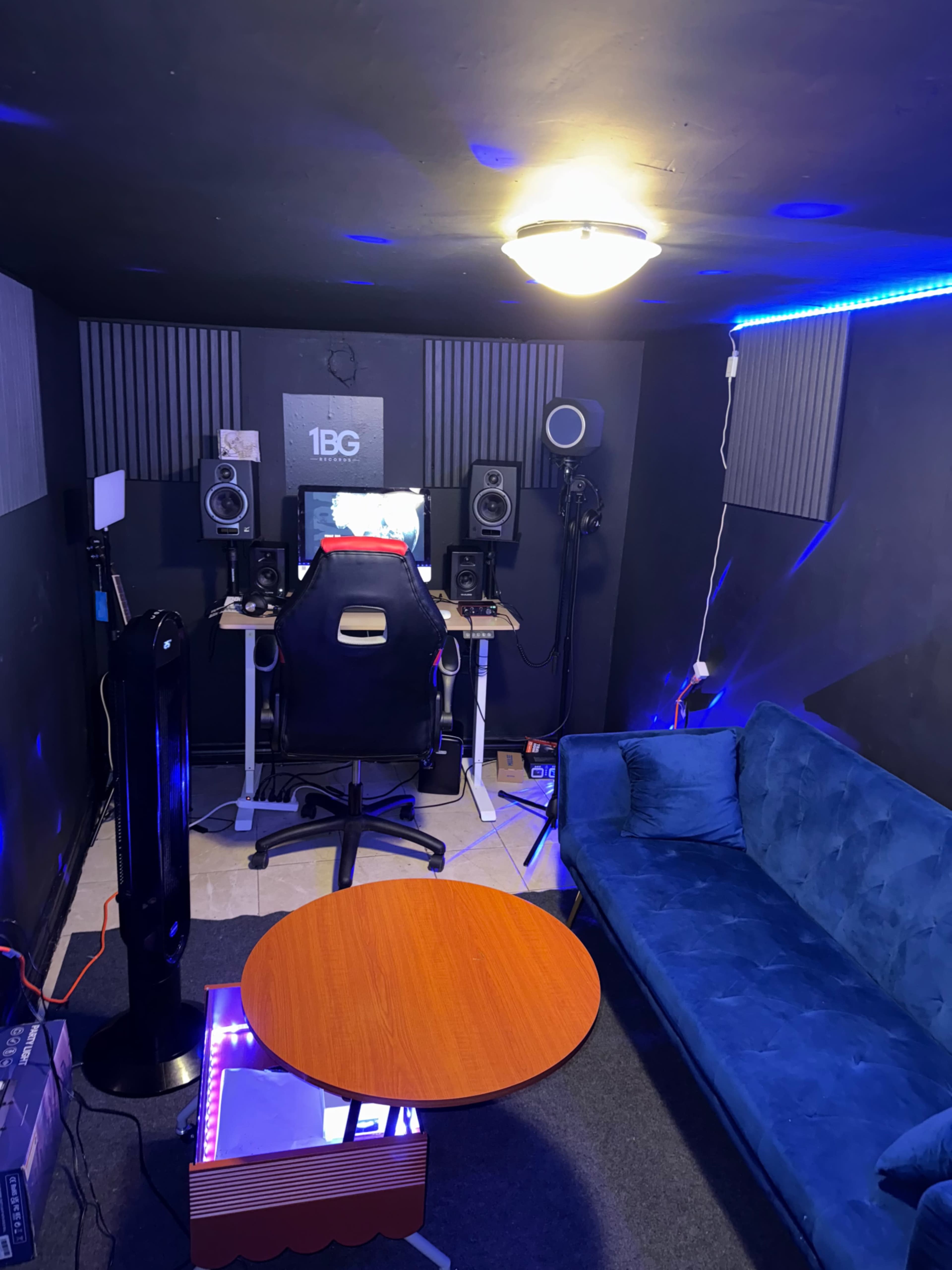 The image shows a small recording studio with a desk, computer, speakers, and a teal couch illuminated by LED lights.
