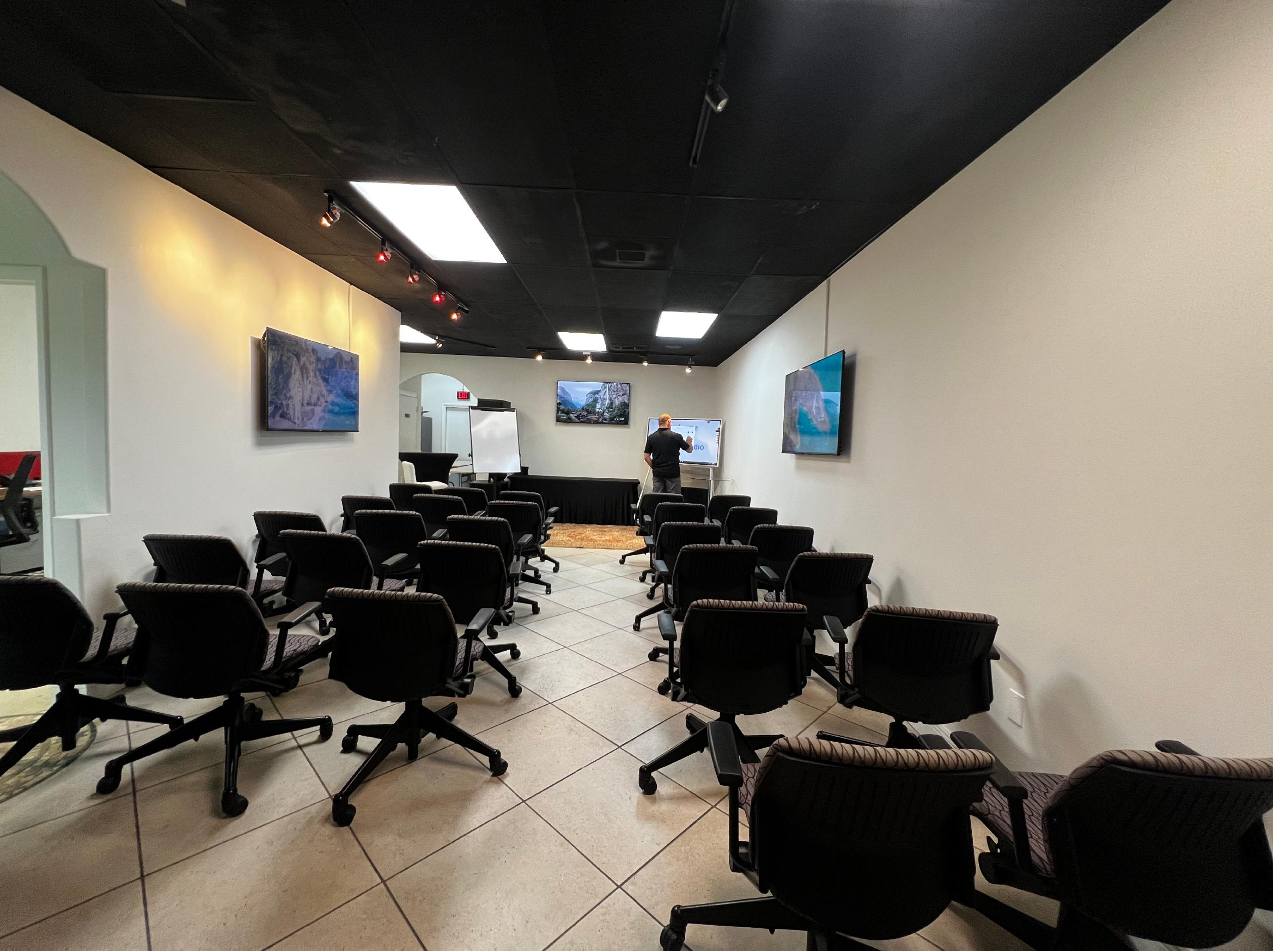 A meeting room is set up with multiple chairs facing a presentation area, where a person is writing on a whiteboard.