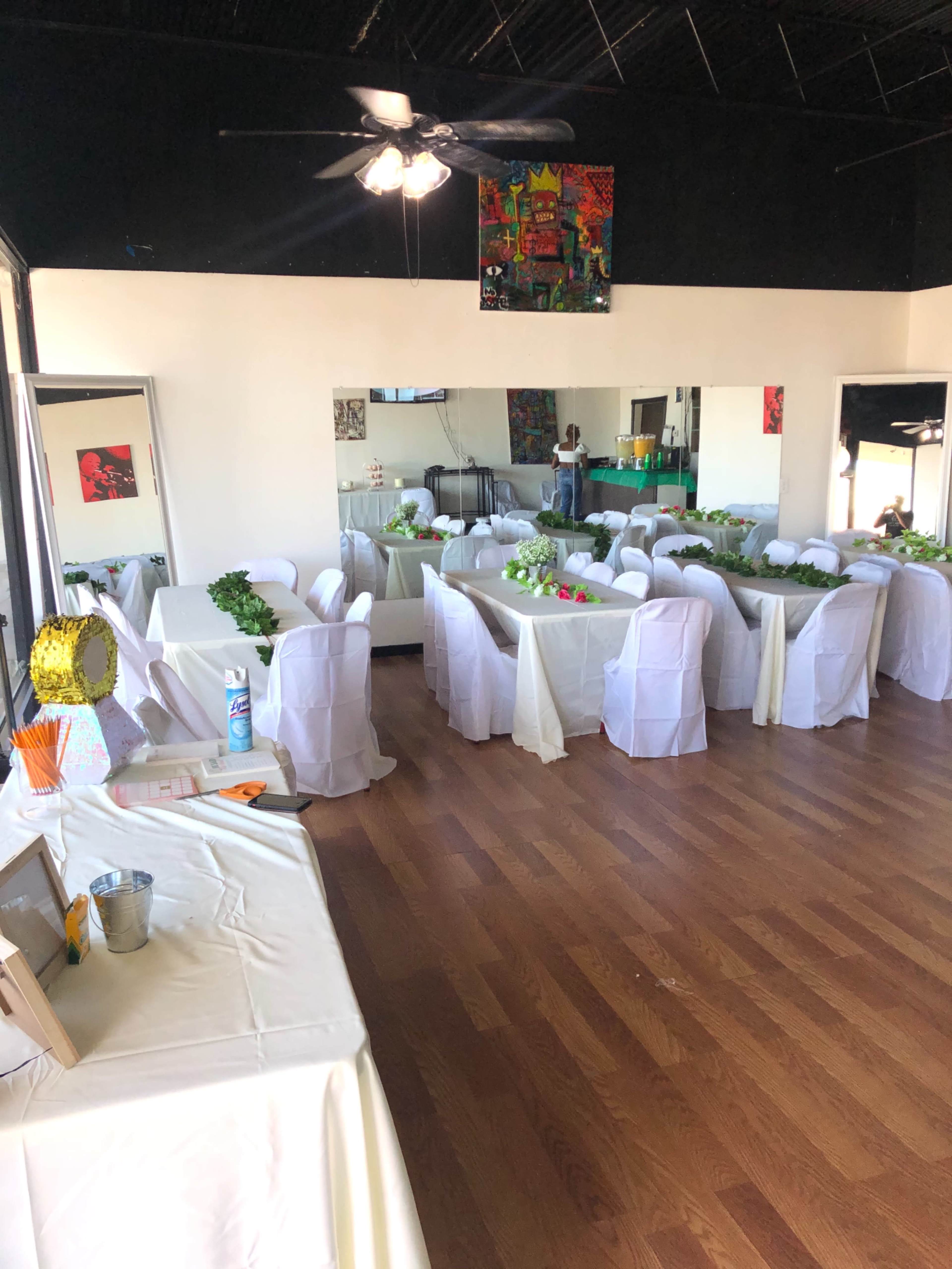 The image shows a spacious event room set up with white tablecloths and chairs, arranged for a gathering with decorations on the tables.