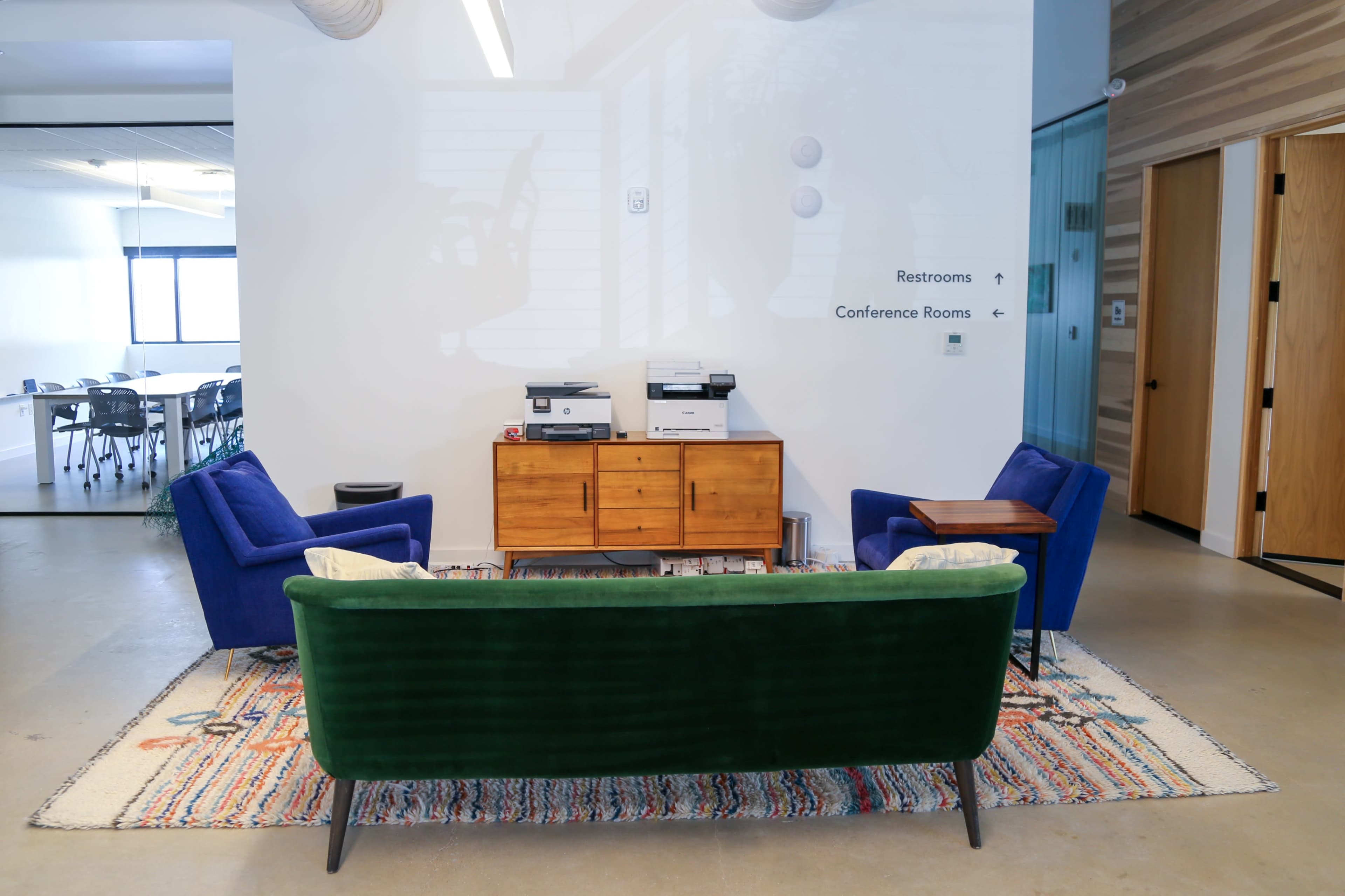 The image shows a modern office space featuring a green velvet sofa, two blue chairs, a wooden cabinet, and printers against a wall with signage indicating restrooms and conference rooms, all situated on a colorful rug.