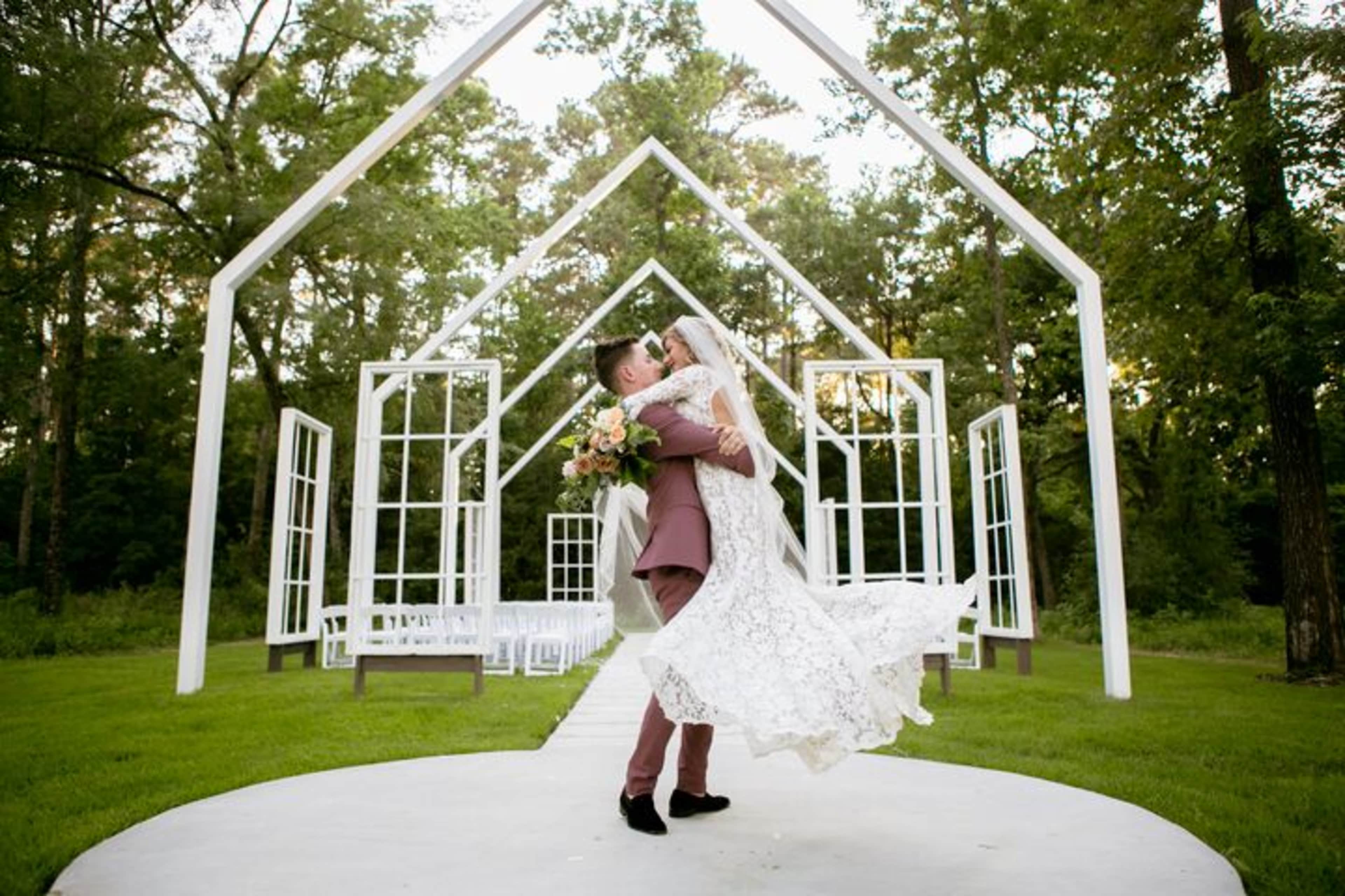 A groom lifts his bride in front of a white-framed wedding altar surrounded by trees.