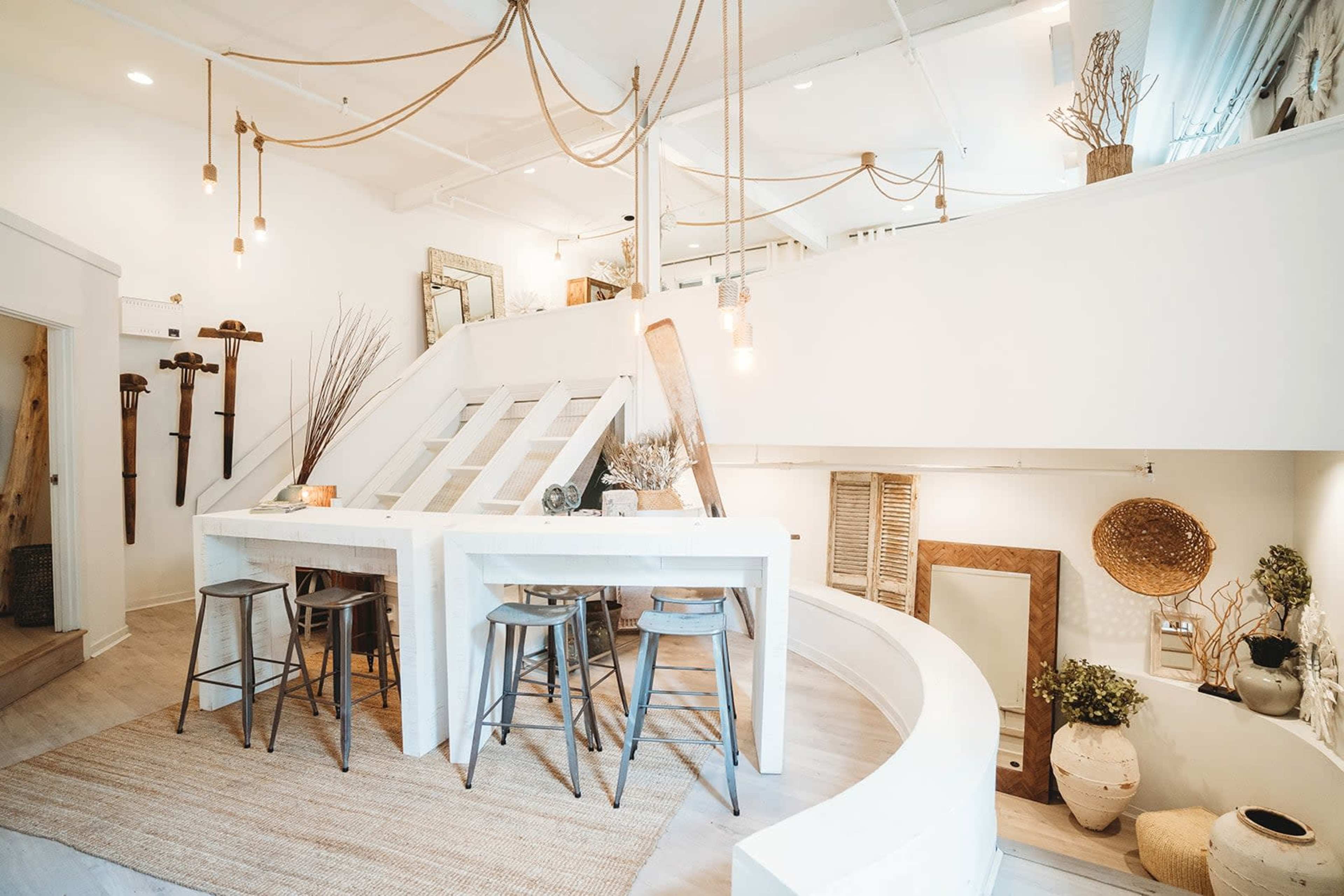 A spacious, modern interior features a light-filled area with a curved table surrounded by metal stools, stairs leading to an upper level, and decor elements like plants and woven baskets.