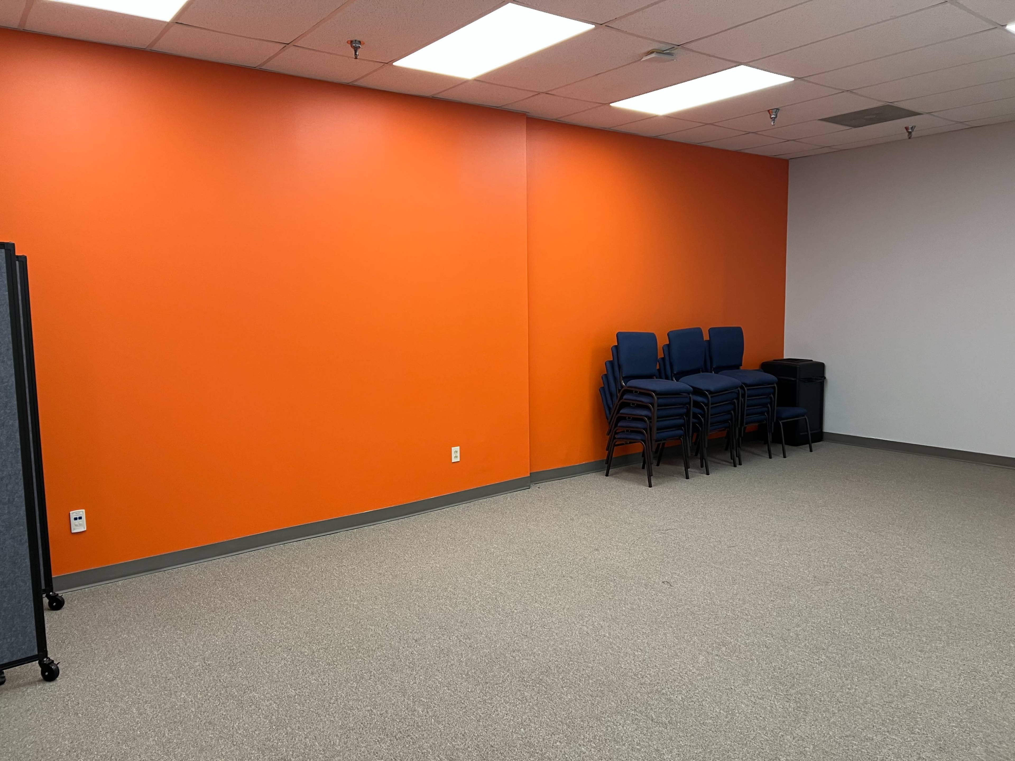 The image shows a sparse room with an orange wall, a stack of blue chairs in the corner, and a trash bin against a gray wall.
