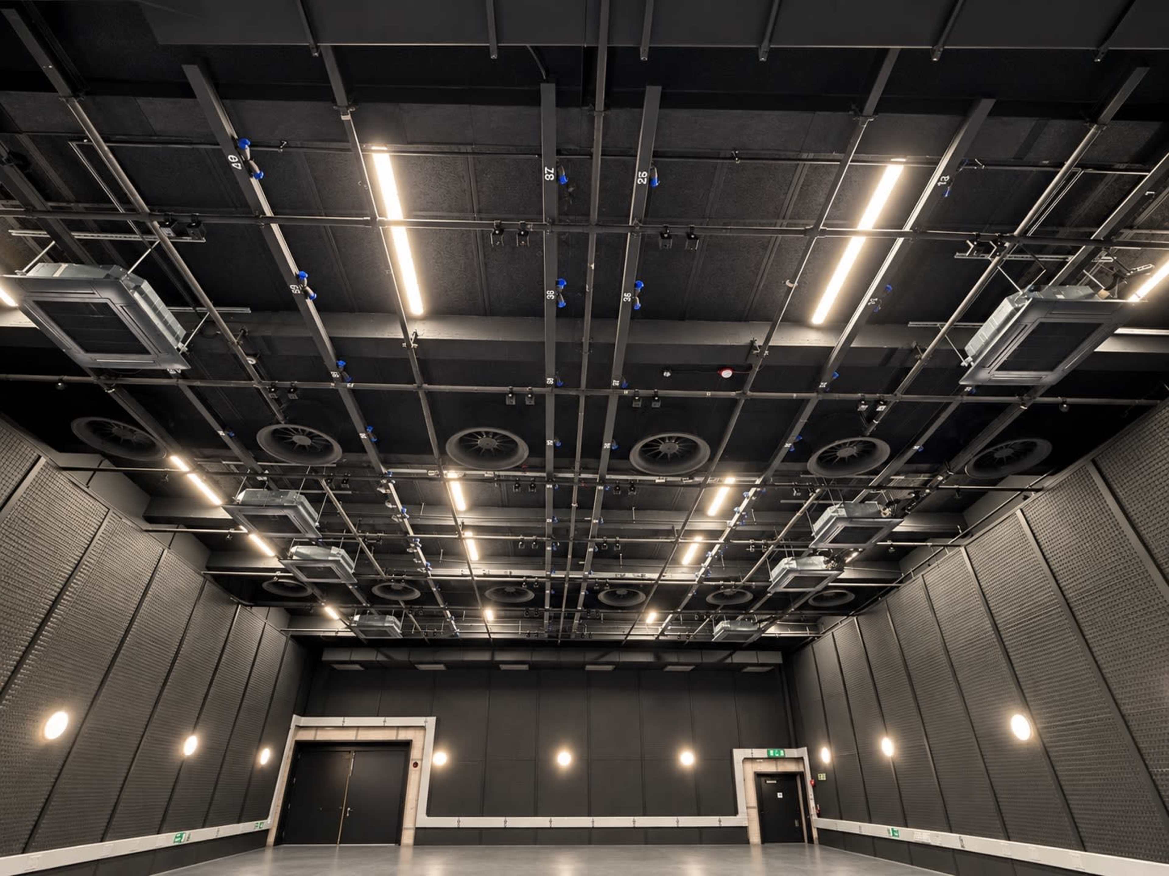 The image shows a spacious indoor room with a high ceiling adorned with ventilation units and overhead lighting.