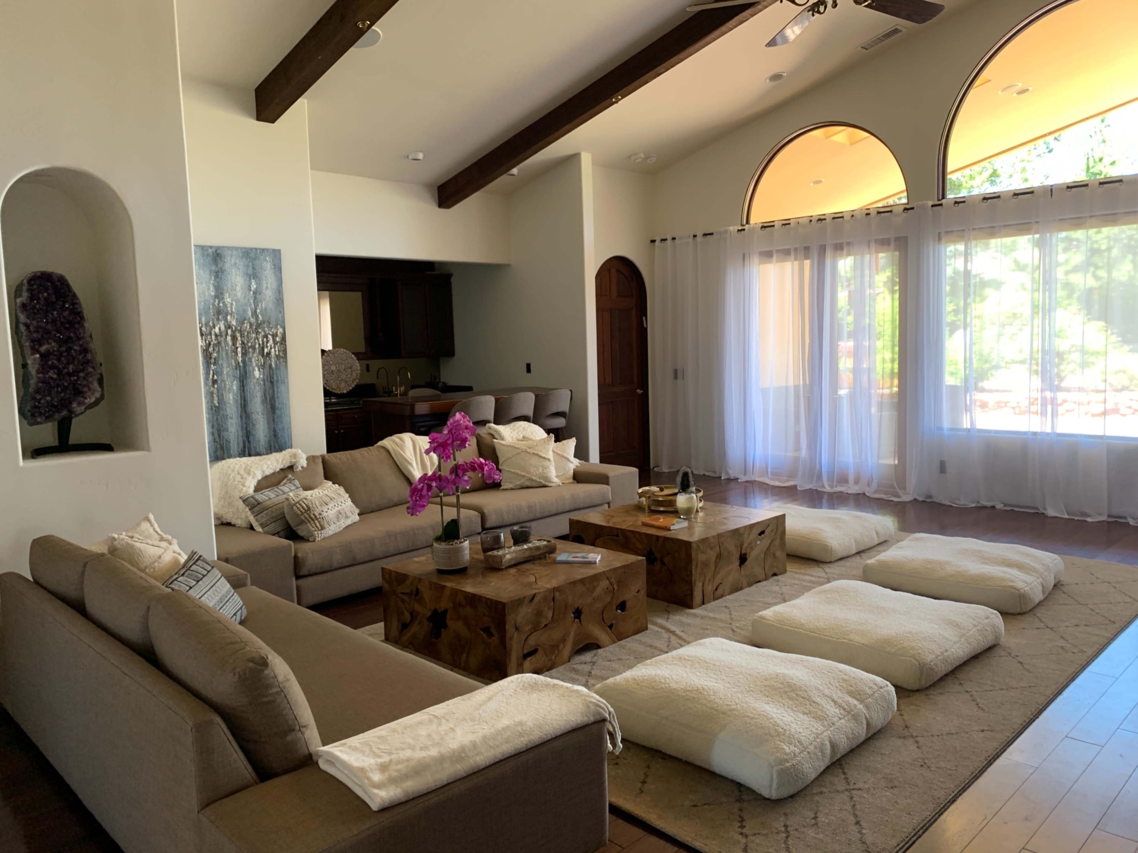 A spacious living room features neutral-toned sofas, wooden tables, and floor cushions, with large windows allowing natural light to enter.