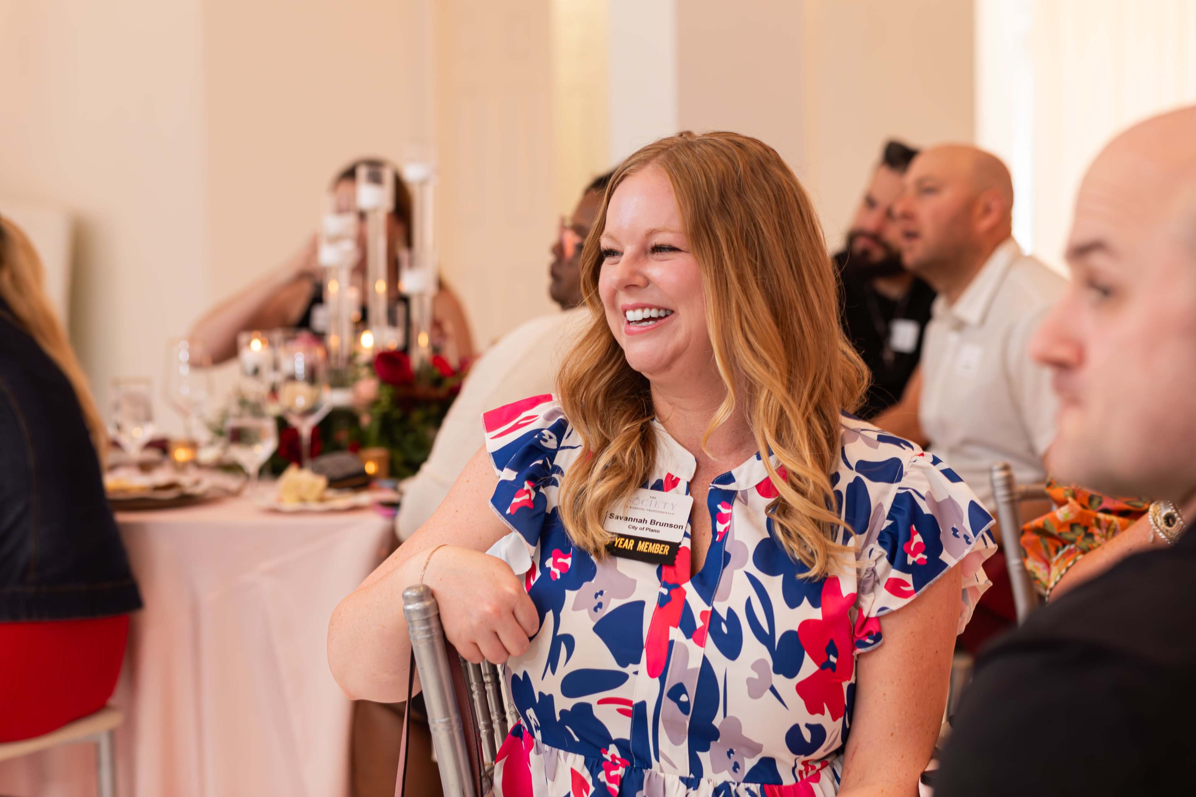 A woman in a colorful dress smiles while seated at a dining table surrounded by other guests at a gathering.