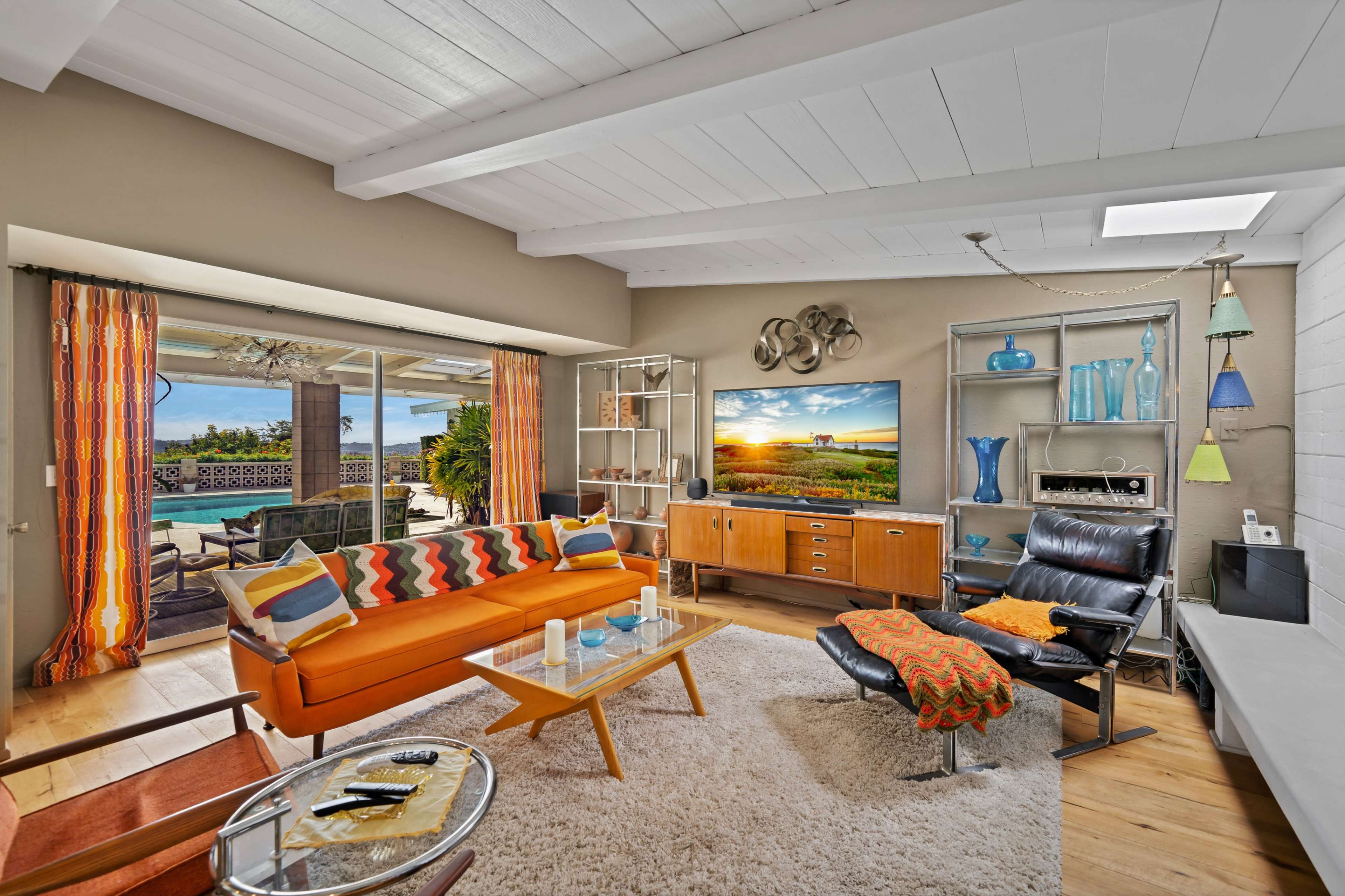 A modern living room features a large orange sofa, a coffee table, a sleek black chair, and a wall-mounted television, all arranged around a brightly lit space with large windows overlooking an outdoor area.