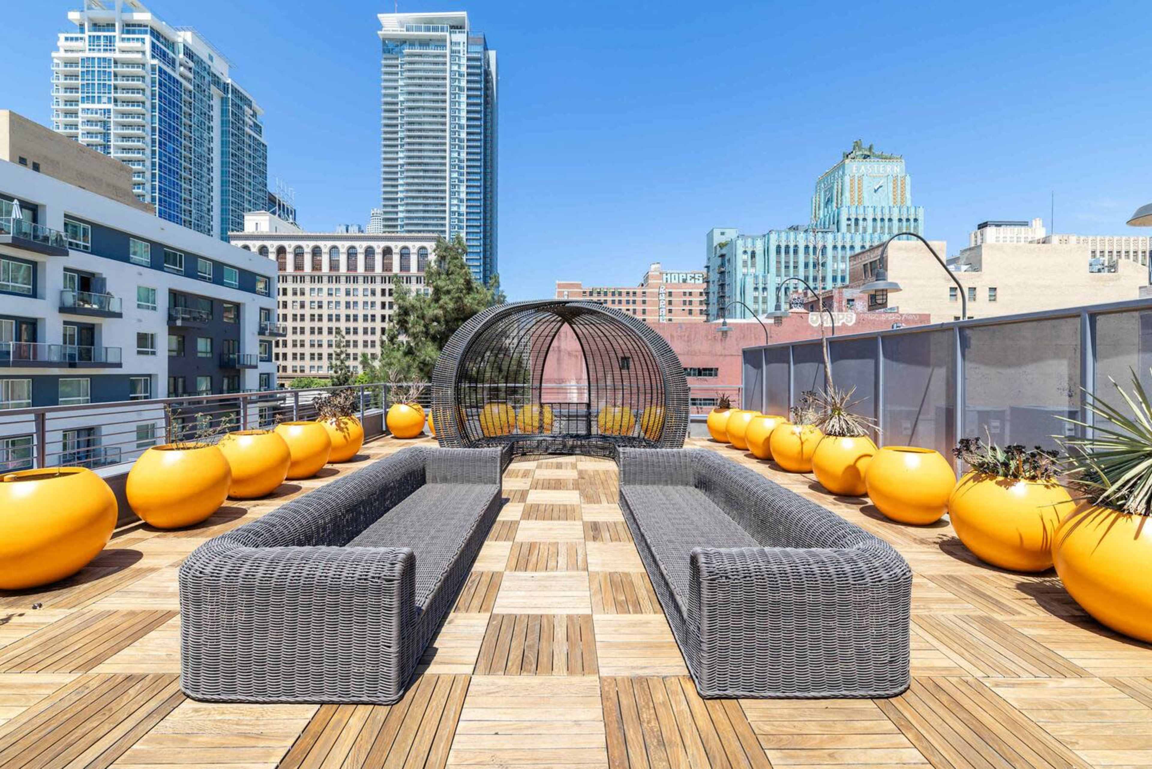 The image shows a rooftop lounge area with gray wicker furniture and large yellow planters, set against a backdrop of modern urban buildings under a clear blue sky.