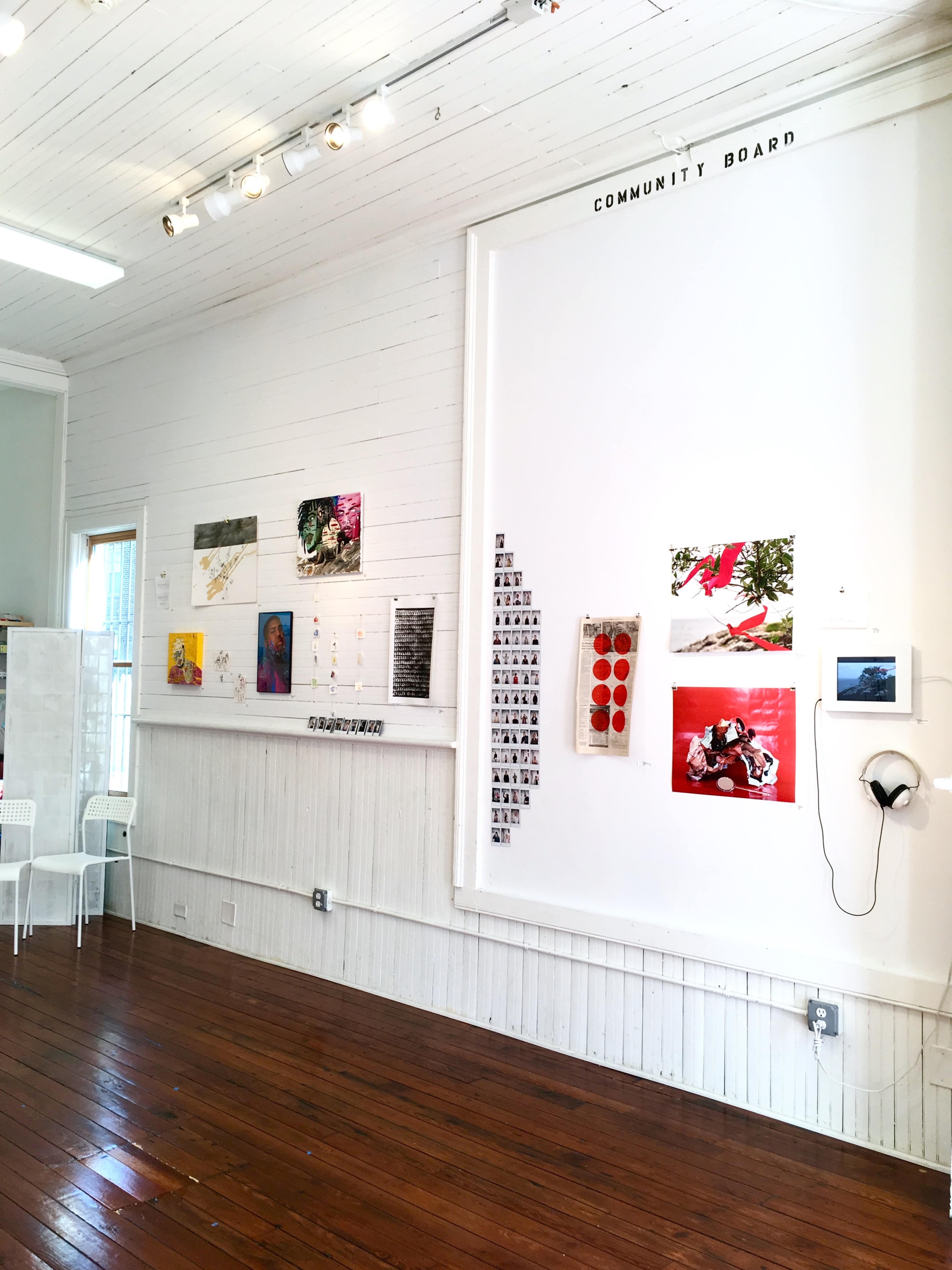 The image shows a gallery wall featuring a variety of artworks and a community board in a well-lit wooden room.