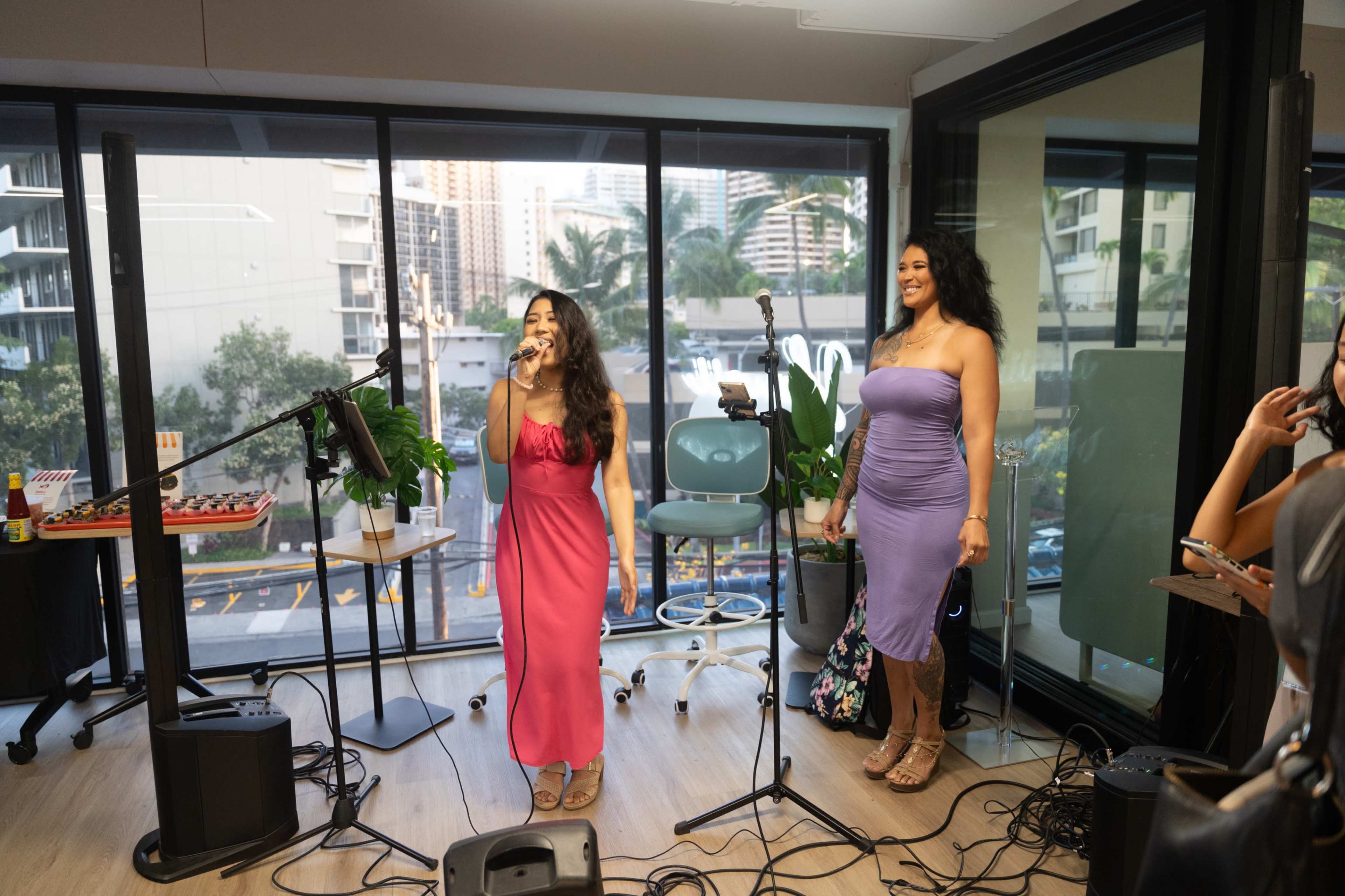 Two women stand in front of microphones in a brightly lit room with large windows, one singing and the other looking on, while musical equipment is set up nearby.