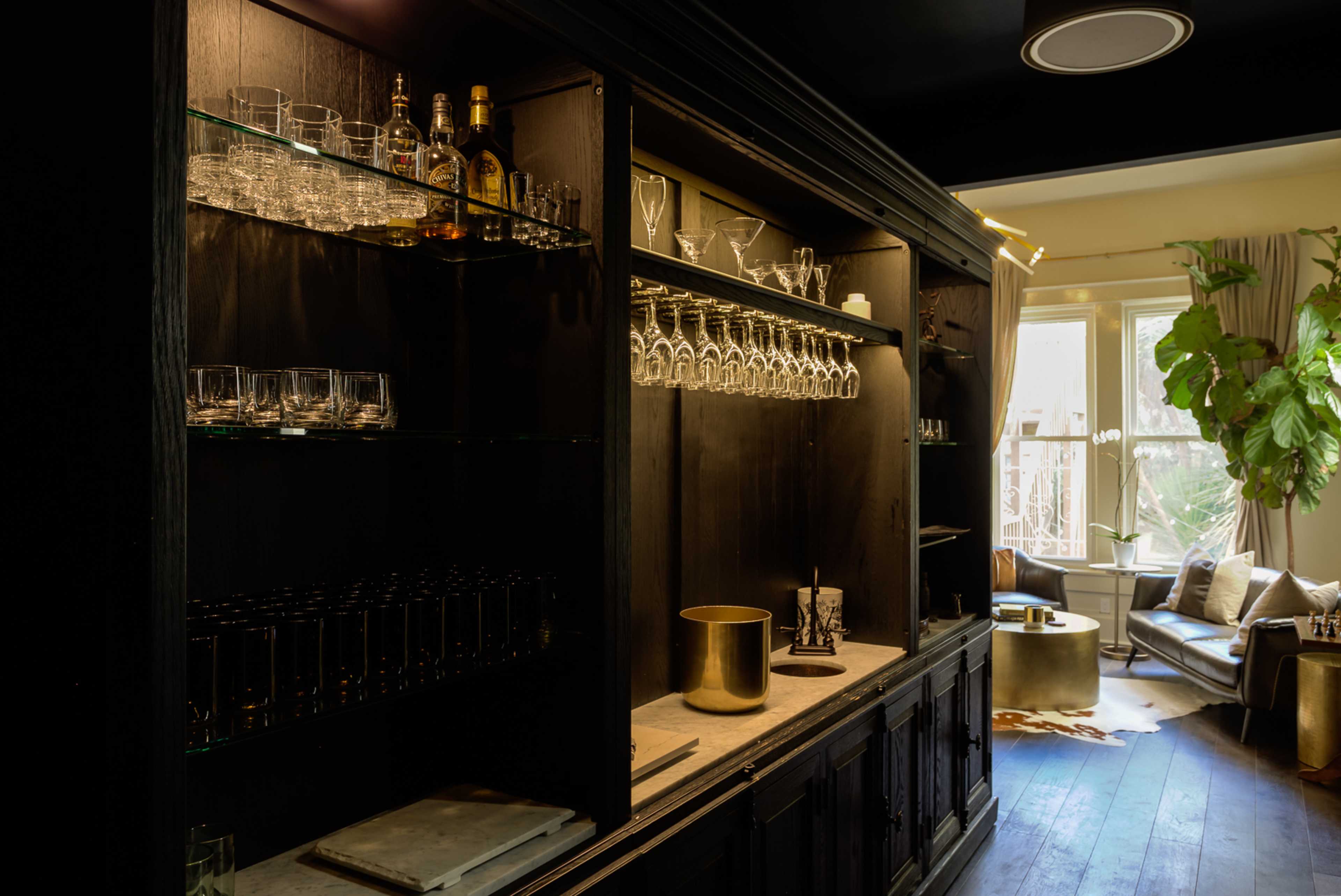 A dark wood bar cabinet with shelves containing various glassware and bottles, adjacent to a well-lit living room with a sofa and a large plant.
