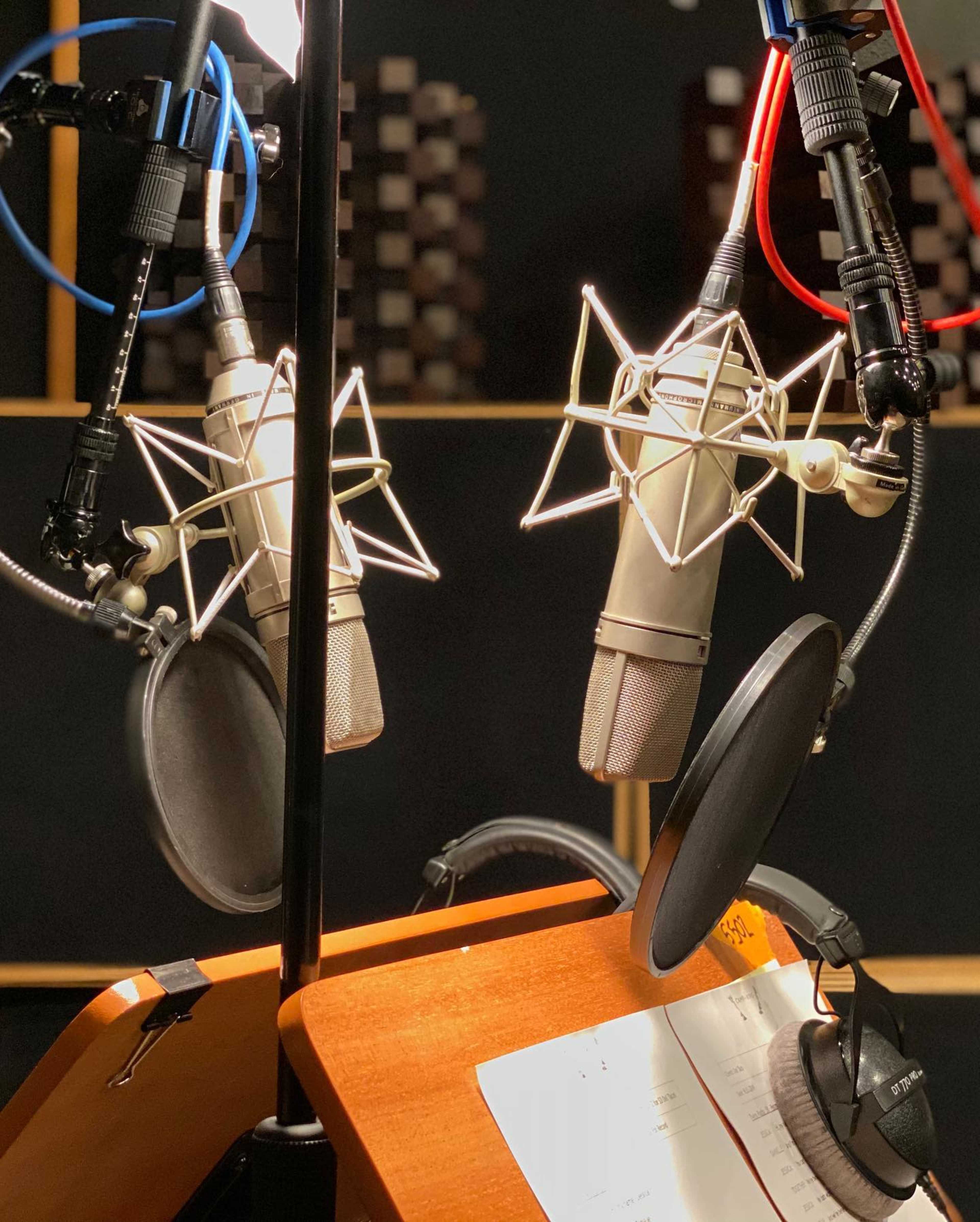 The image shows two studio microphones positioned on a stand in a recording booth with a music score on a wooden surface.