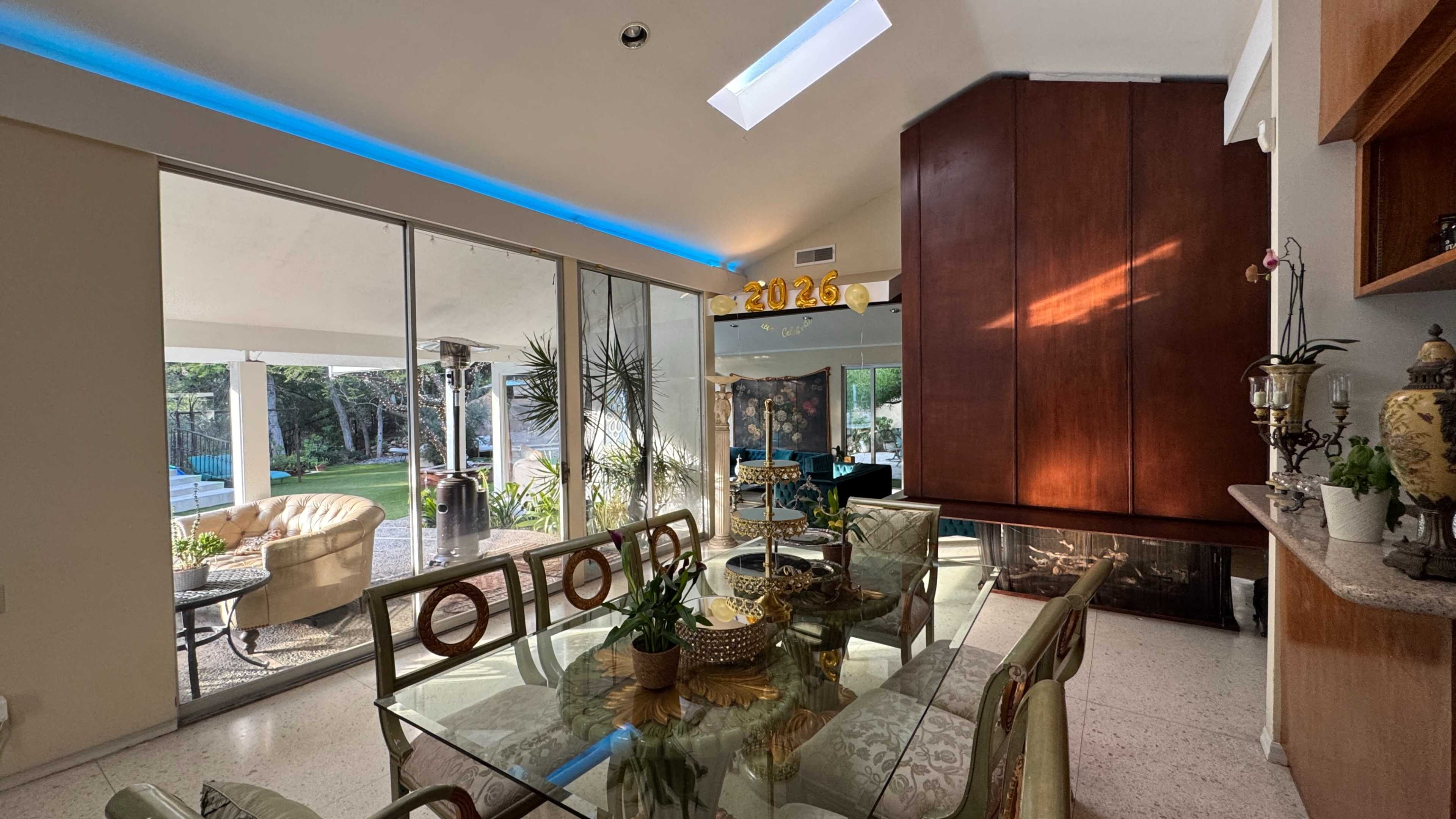 A dining area featuring a glass table with ornate chairs, large windows overlooking a garden, and a wooden accent wall with a modern fireplace.