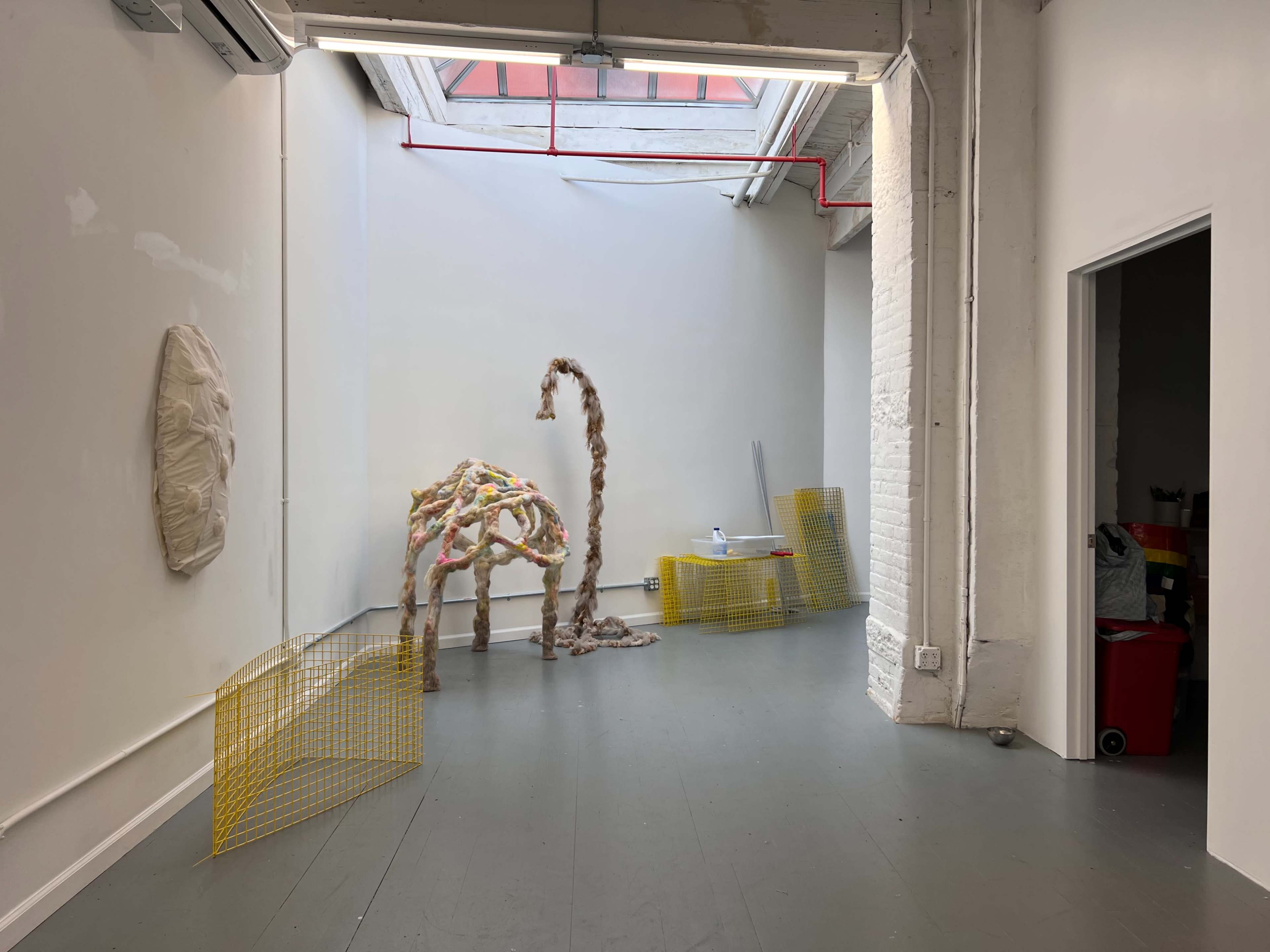 An art gallery space featuring a textured sculpture resembling an animal, alongside yellow mesh fencing and various articles against the walls.