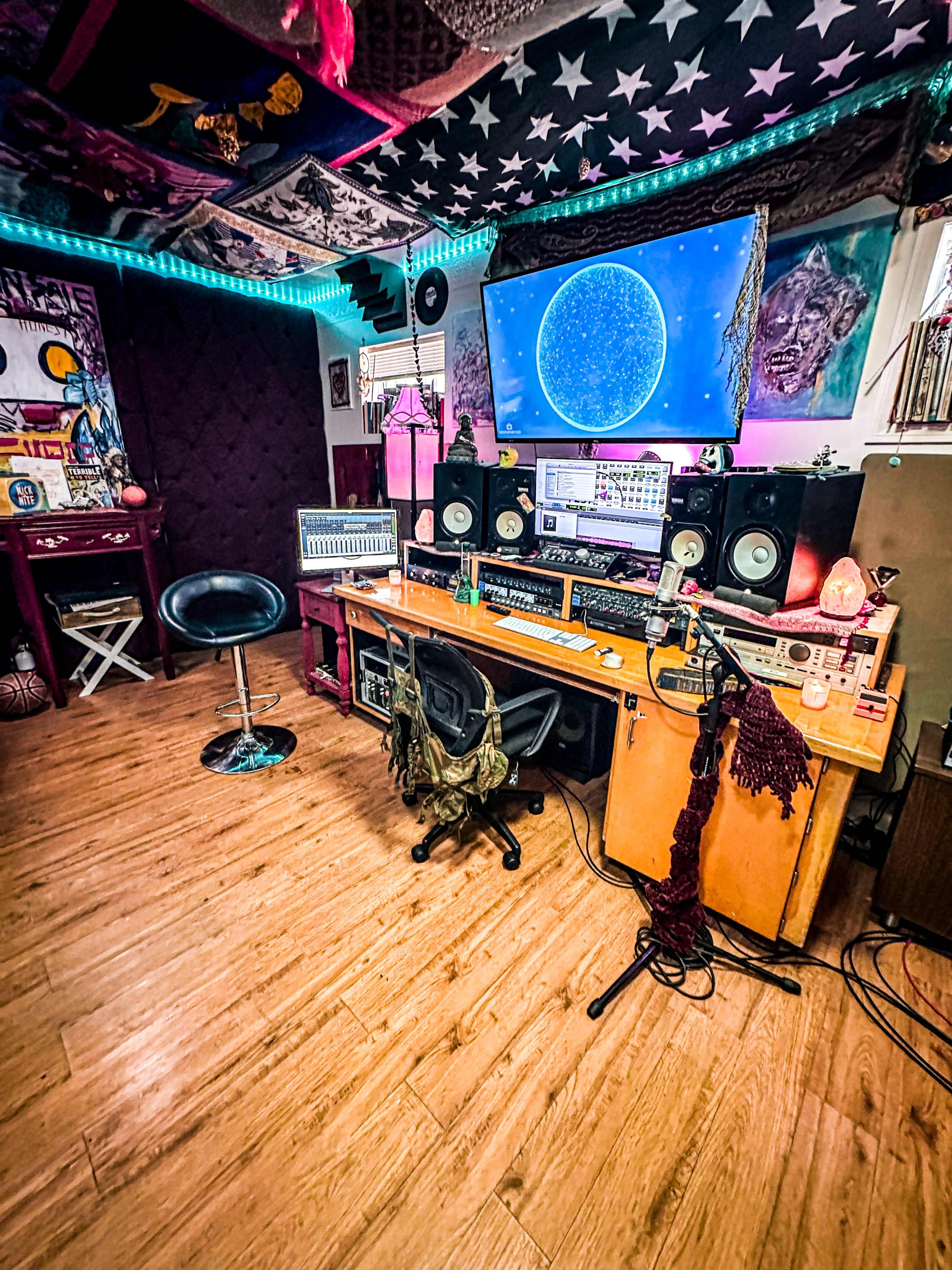 The image shows a home music studio featuring a large monitor, audio equipment, a mixing console, and colorful lighting.