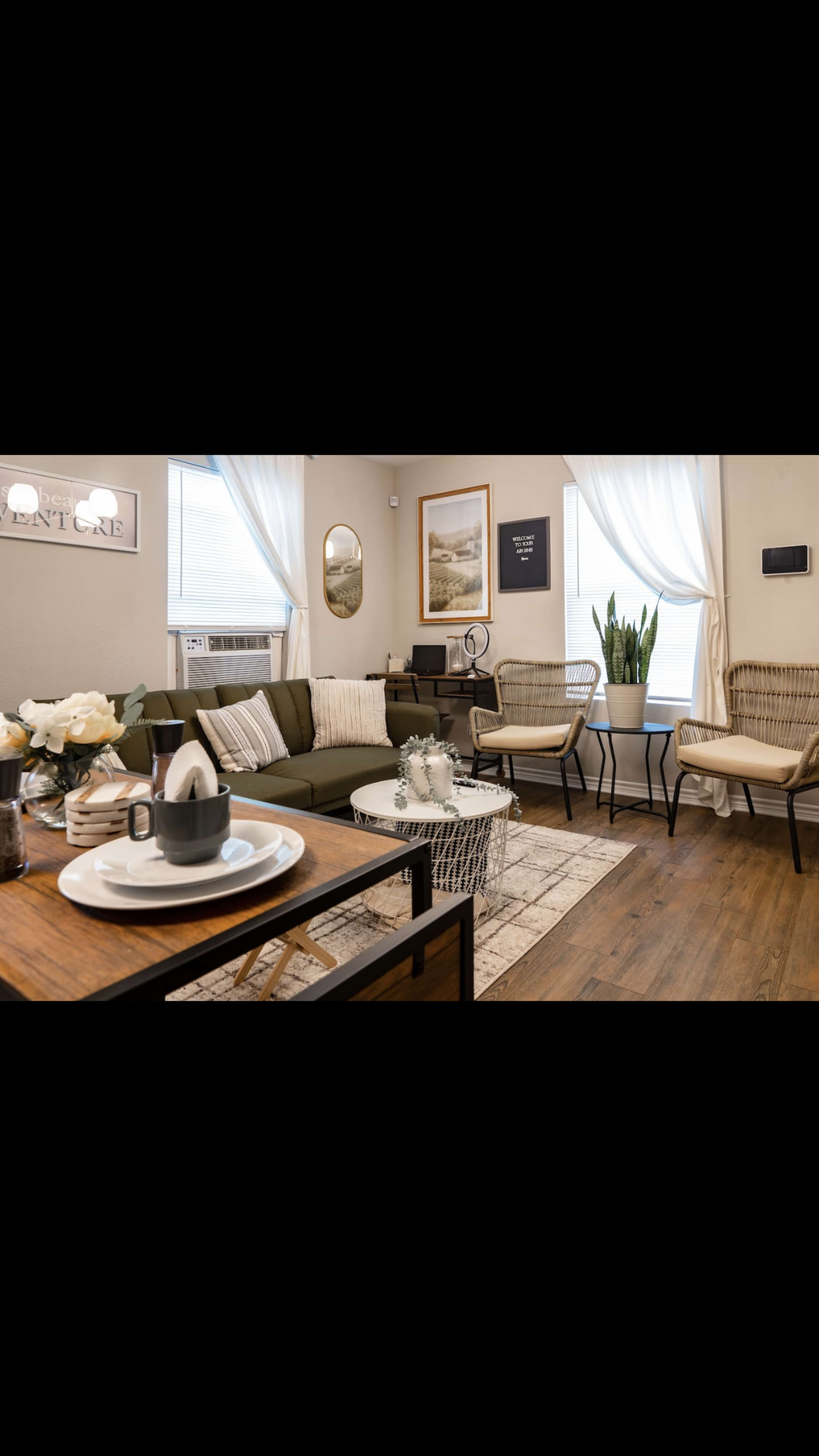 A modern living room features a green sectional sofa, a wooden coffee table with decorative items, and two rattan chairs beside large windows with sheer curtains.