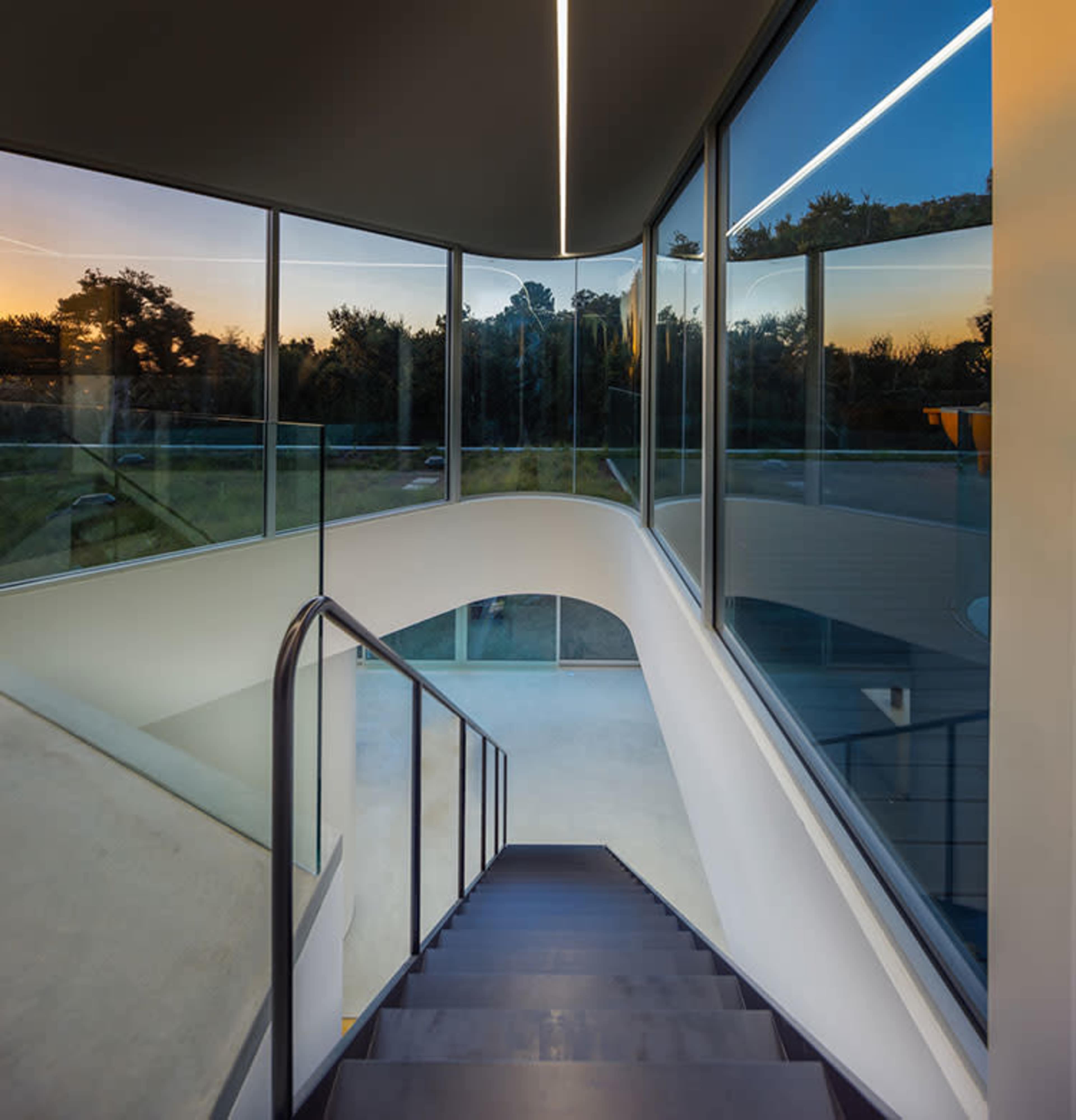 A modern staircase with a sleek railing leads down to a brightly lit space surrounded by large glass windows.