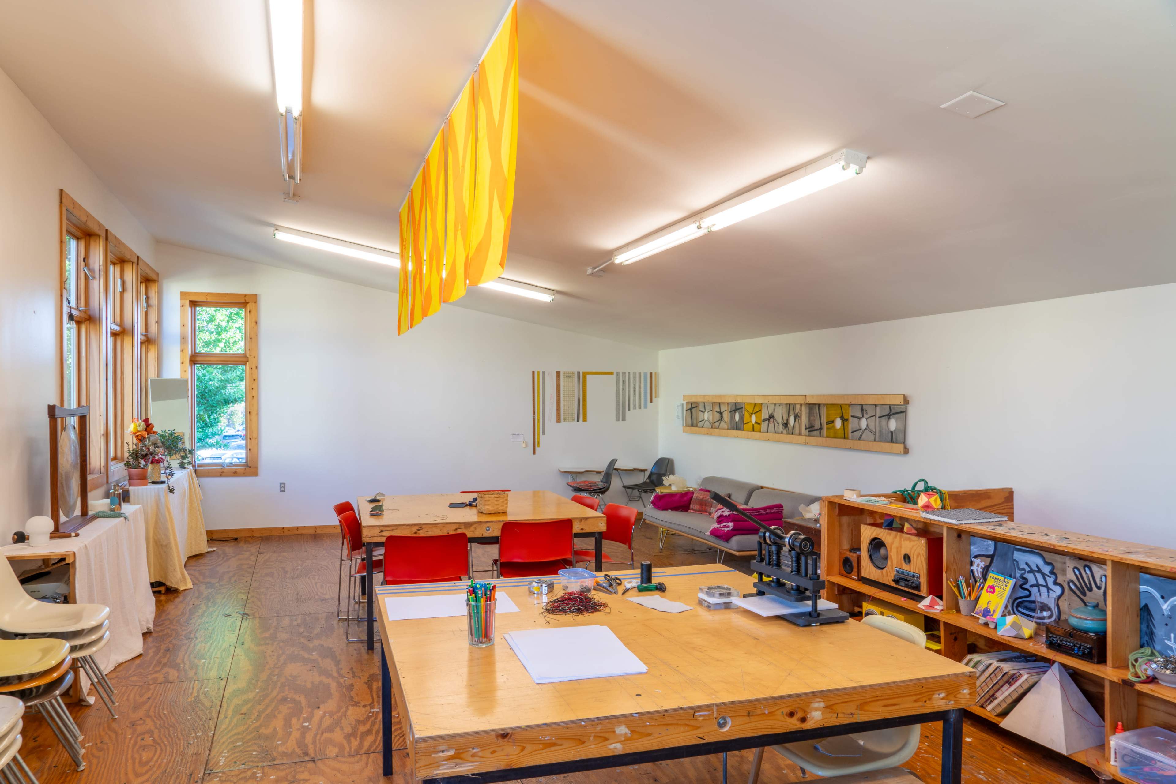 An art studio with wooden tables, chairs, and bright lighting, featuring various art supplies and a decorative curtain hanging from the ceiling.