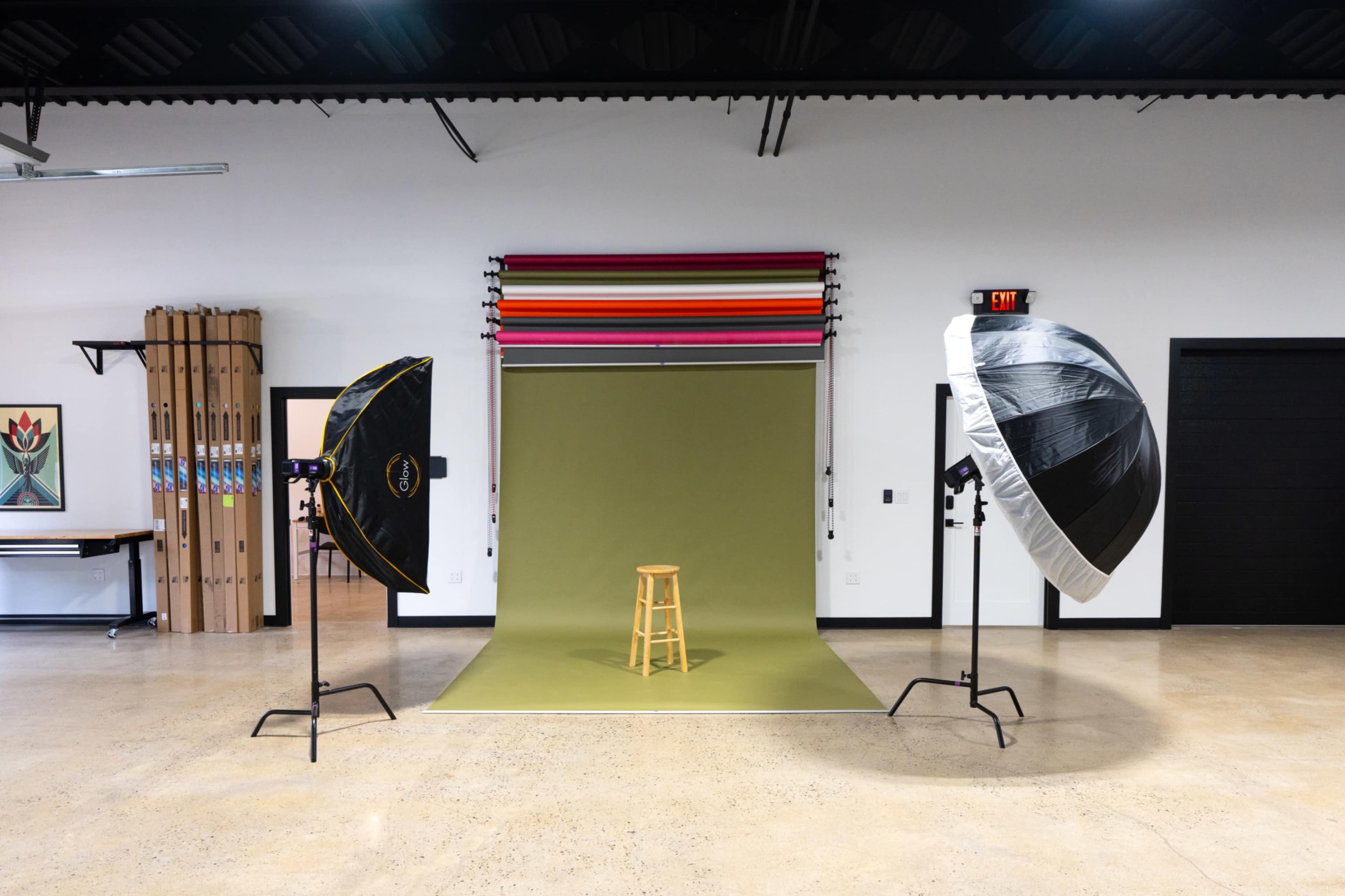 The image shows a photography studio setup with two softbox lights, a green backdrop, and a wooden stool positioned in front of it.