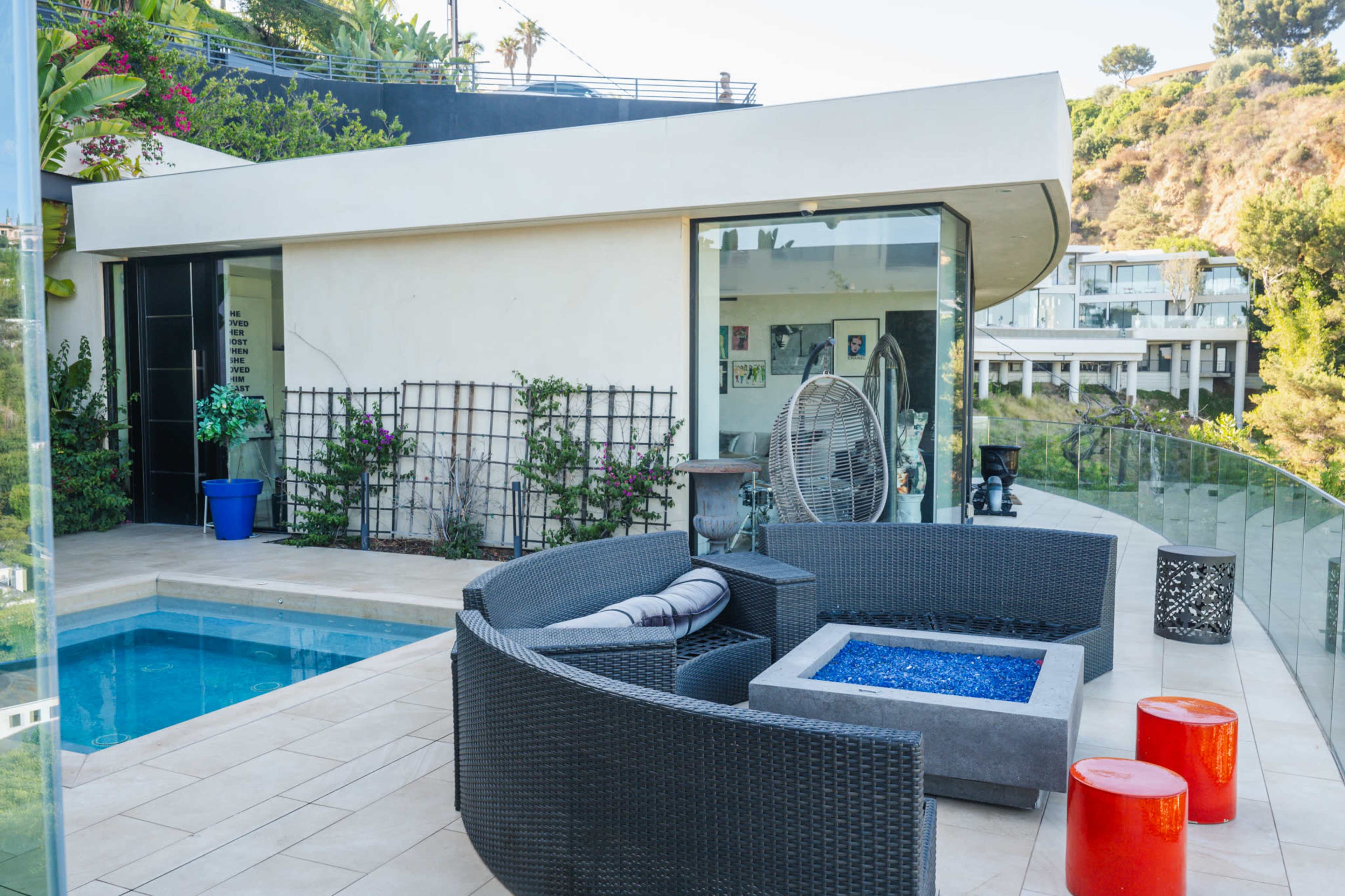 The image shows a modern outdoor patio area with a pool, a seating arrangement featuring dark wicker furniture, and a glass railing overlooking a hillside.