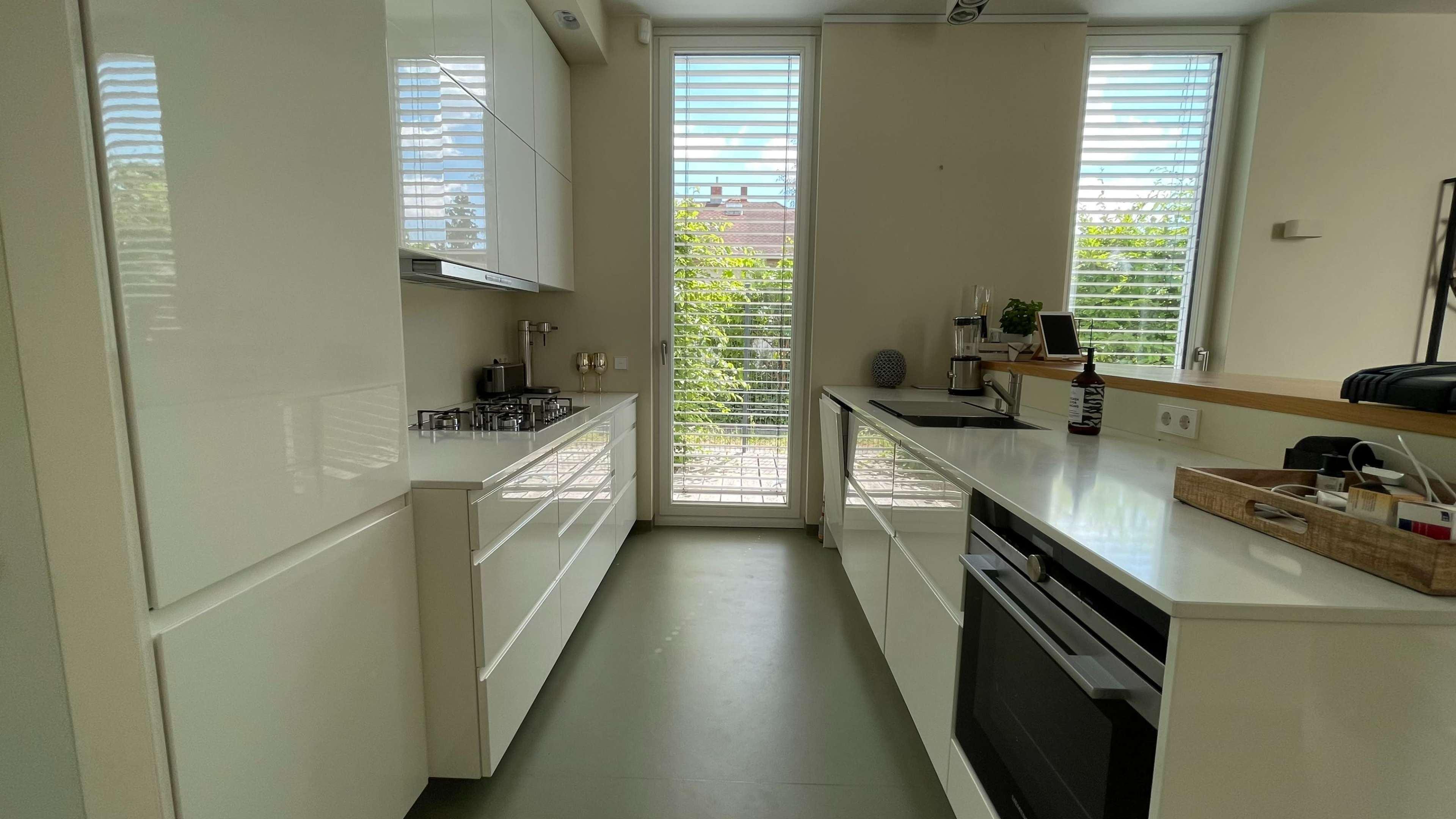 A modern kitchen features white cabinets, a sleek countertop, and large windows with slatted blinds overlooking a garden.