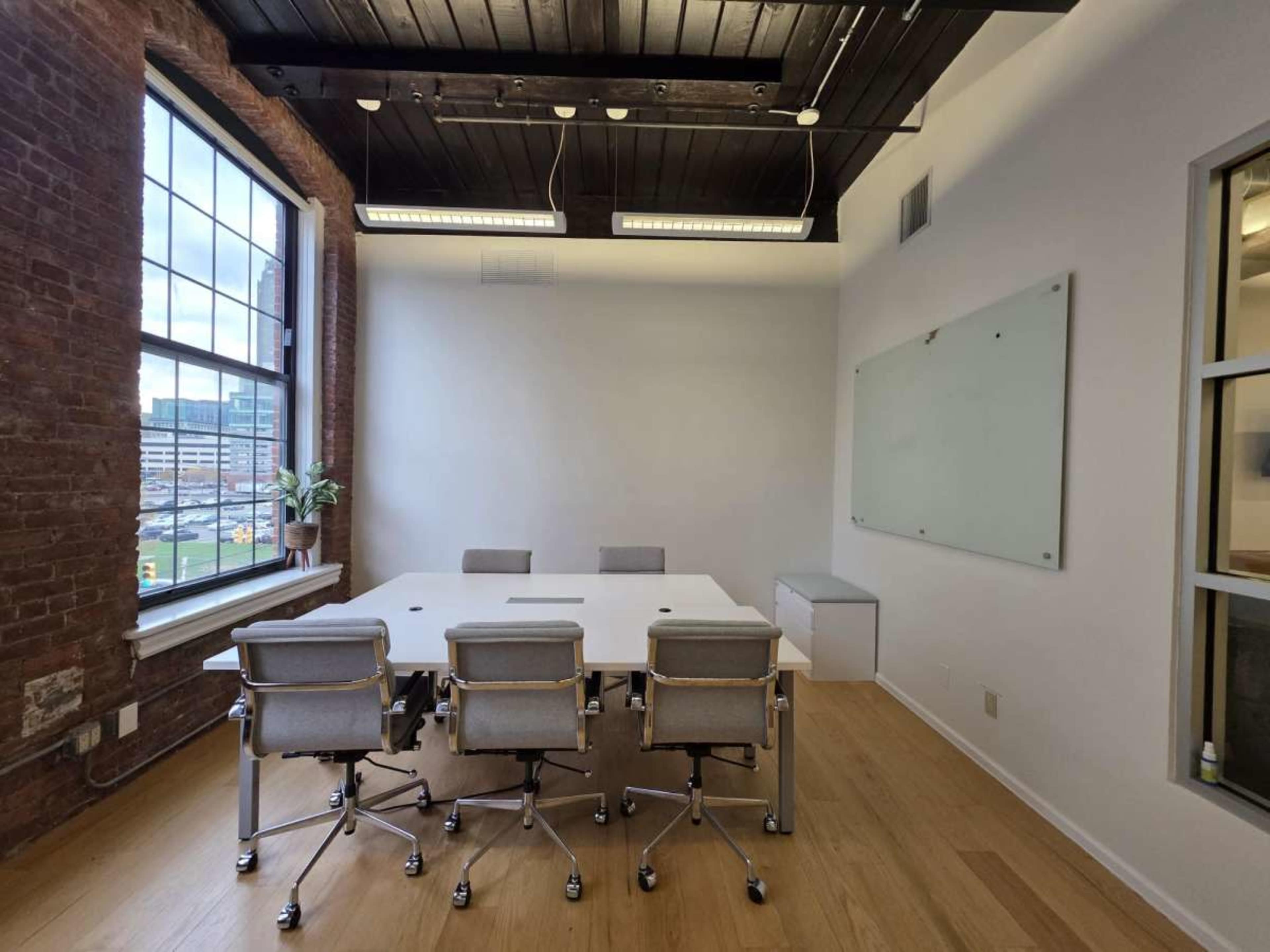 The image shows a modern conference room with a large white table surrounded by several ergonomic chairs, a whiteboard on the wall, and large windows revealing an outdoor view.