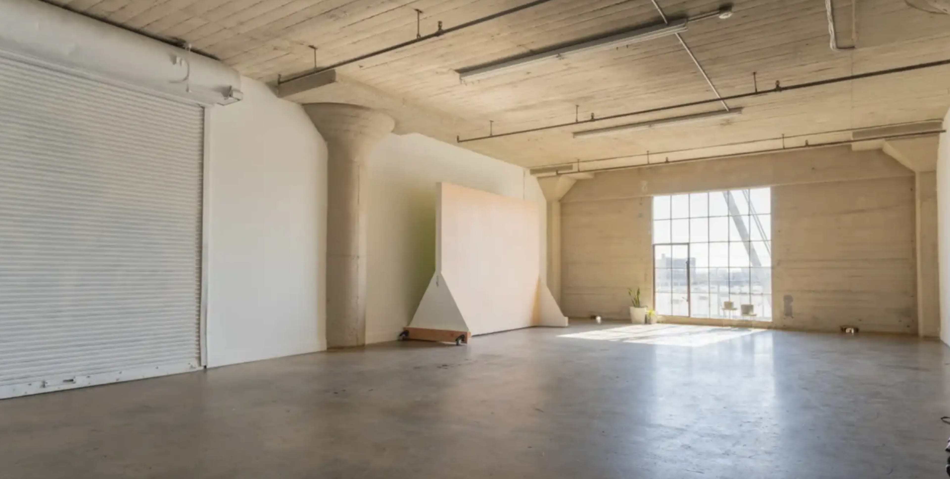 The image shows a spacious, unfinished industrial interior with a large window, a concrete floor, and a minimalist setup featuring a tall wall and a support column.