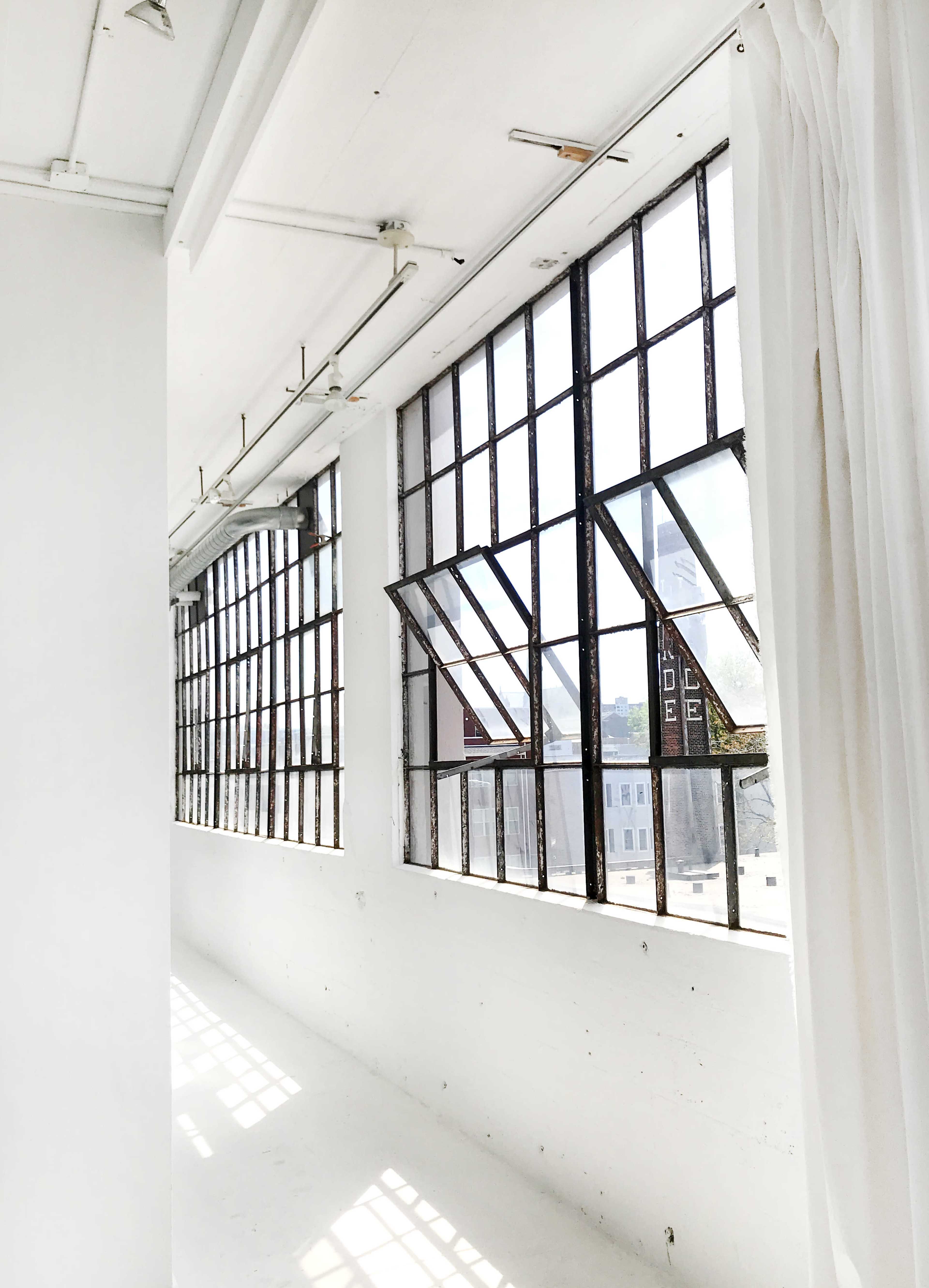 The image shows a bright, open room with large, industrial-style windows that are partially opened, allowing natural light to illuminate the white walls and floor.