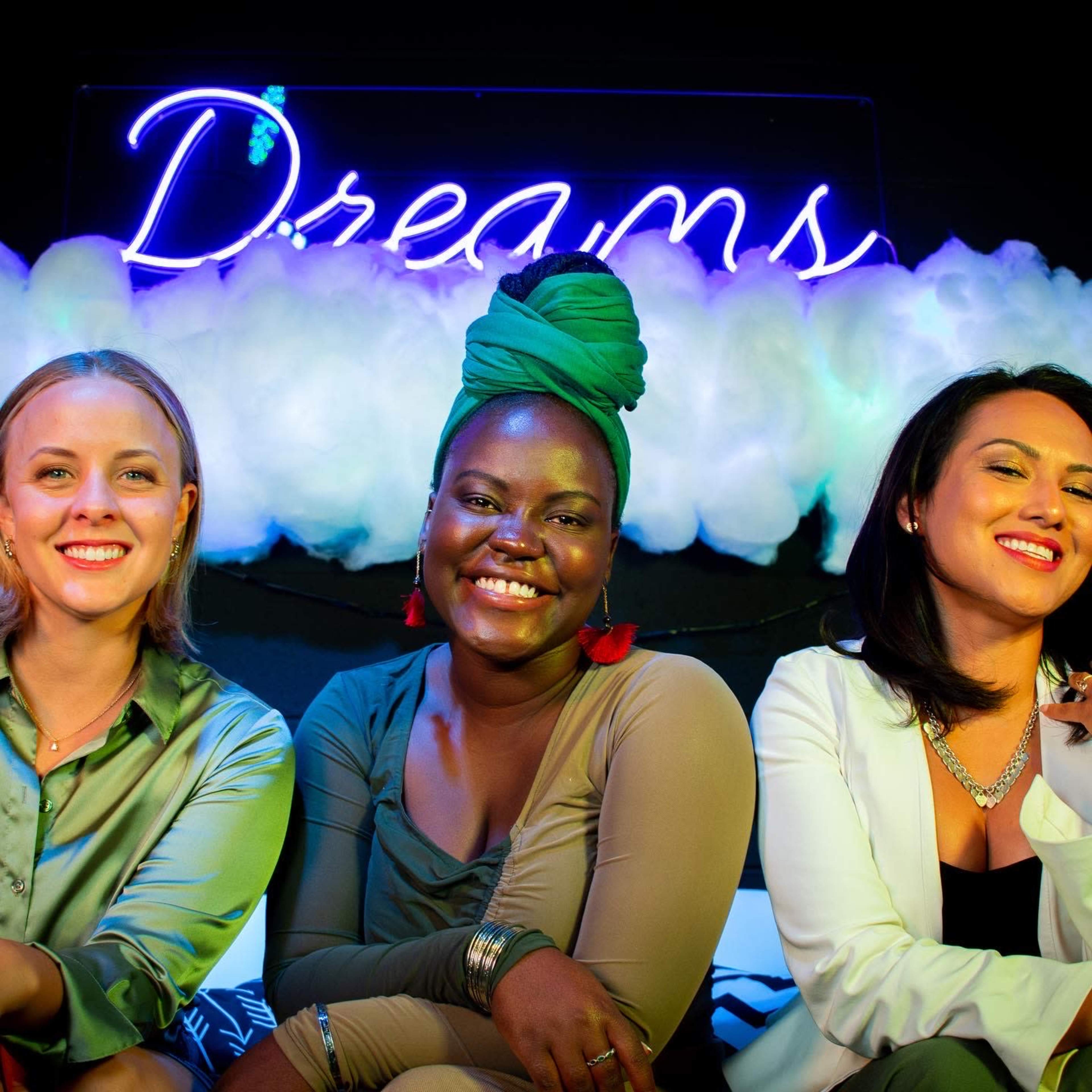 Three women sit closely together, smiling in front of a neon sign that reads "Dreams," with a backdrop of fluffy white clouds.
