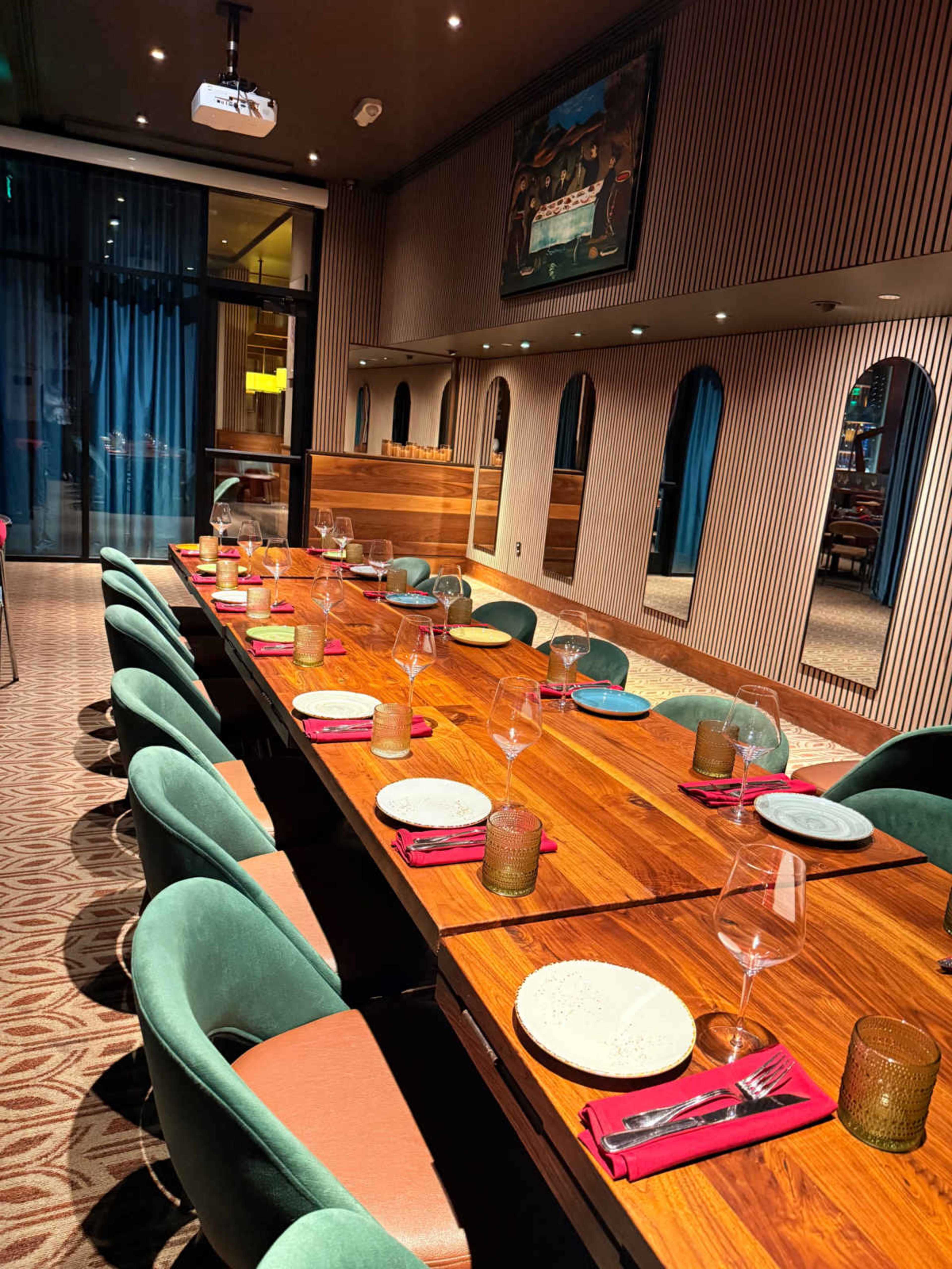 The image shows a long dining table set with plates, utensils, and glasses in a modern restaurant interior, featuring a decor with wooden accents and blue curtains.
