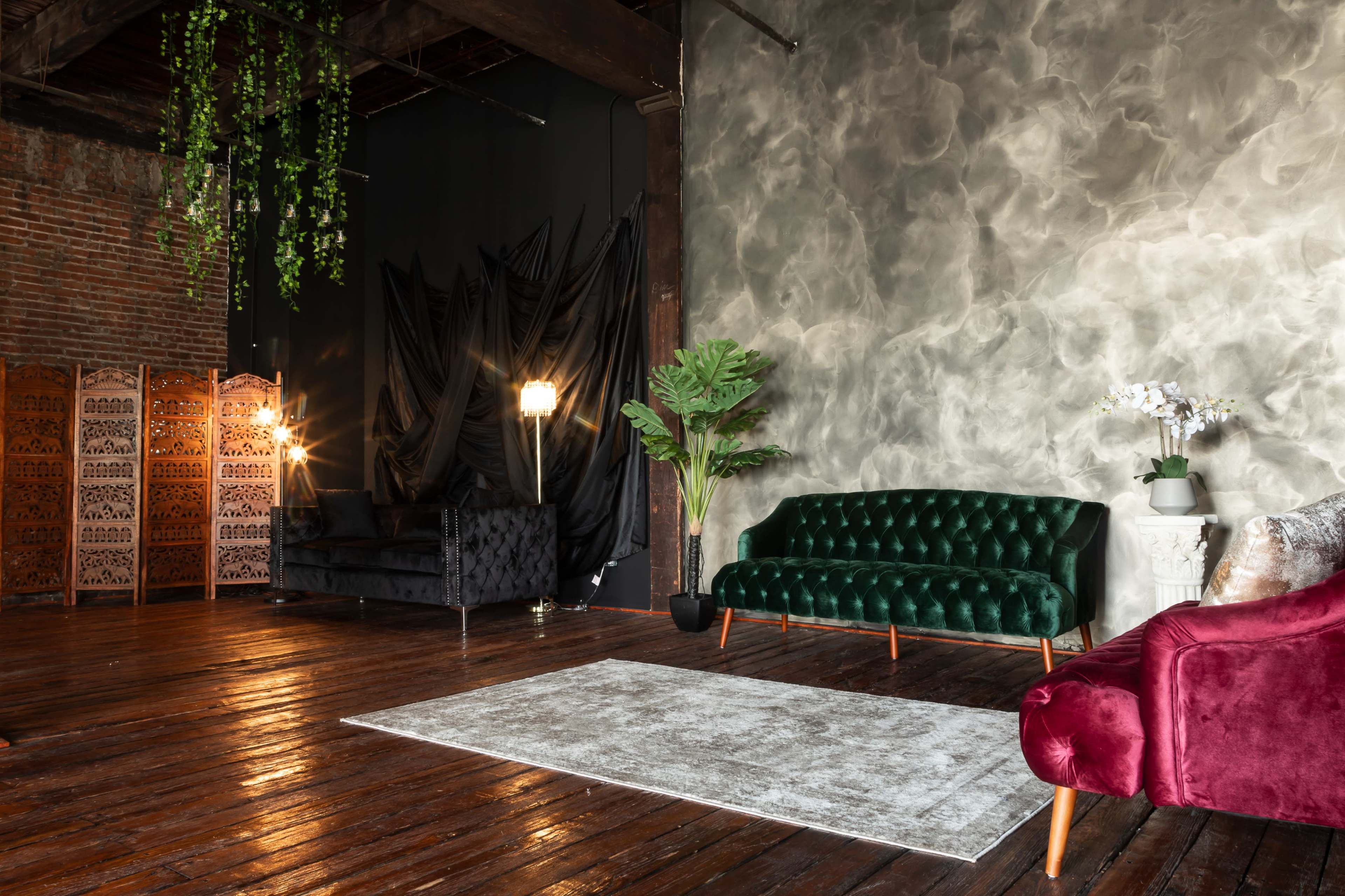 A stylish interior setting with a dark green velvet couch, a burgundy velvet couch, and a large area rug on polished wooden floors, against a textured gray wall and brick background.