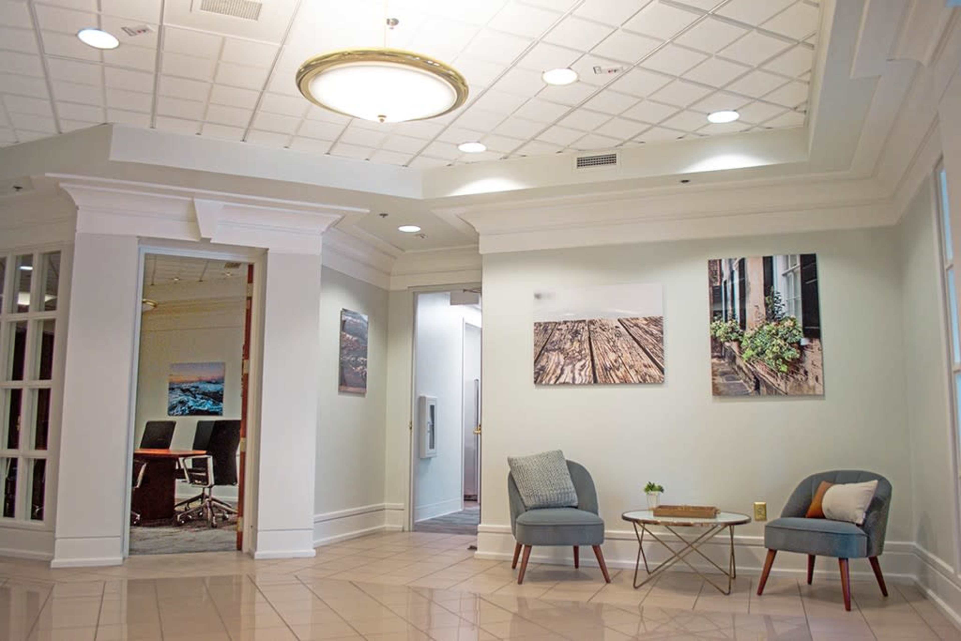 The image shows a well-lit office lobby area featuring two chairs and a small table, with artwork on the walls and a conference room visible in the background.