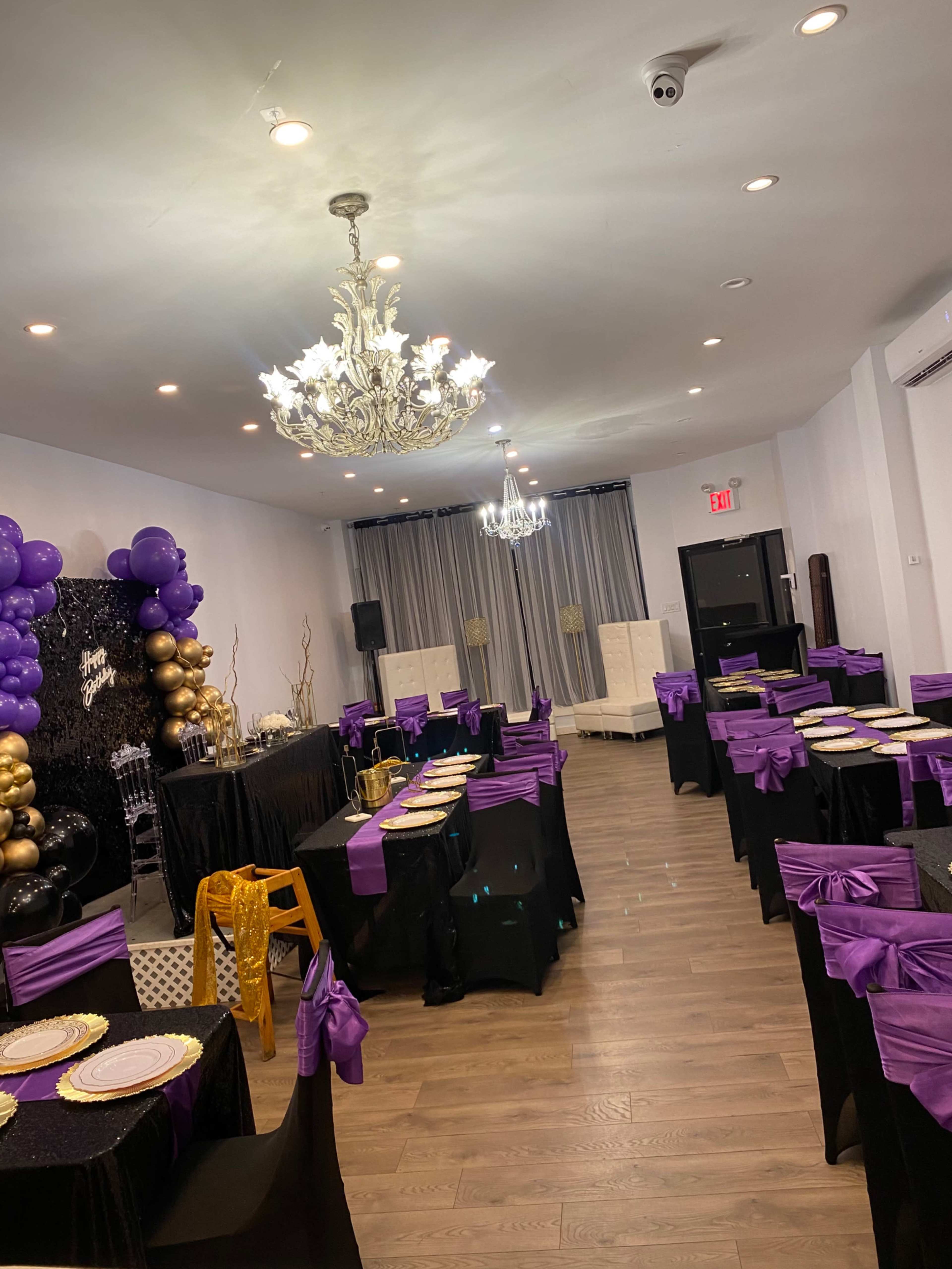 A banquet hall arranged with tables covered in black cloth and purple accents, featuring a decorative backdrop with balloons and a chandelier overhead.