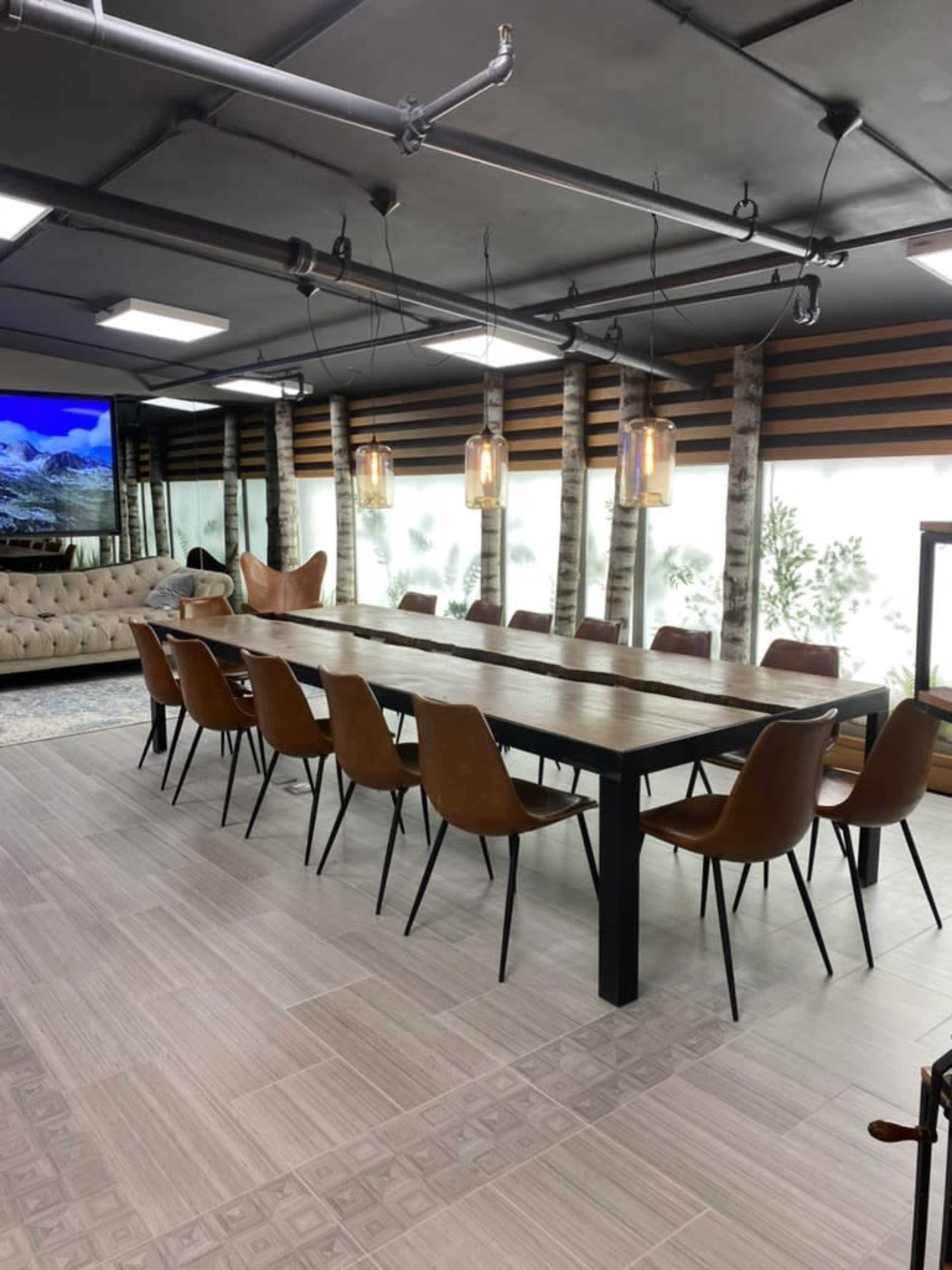 The image shows a modern dining area with a long wooden table surrounded by leather chairs, illuminated by pendant lights, and featuring large windows with plants visible outside.