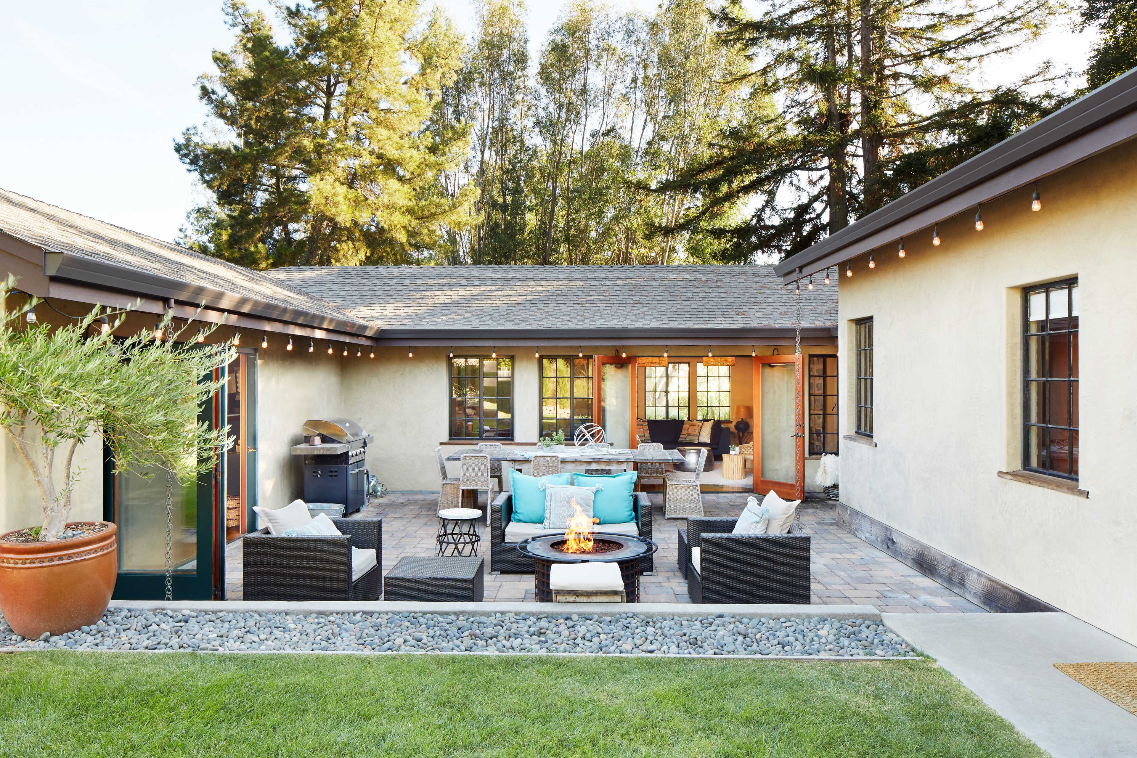 The image shows a backyard patio featuring a fire pit surrounded by seating, a barbecue grill, and string lights overhead against a backdrop of trees.