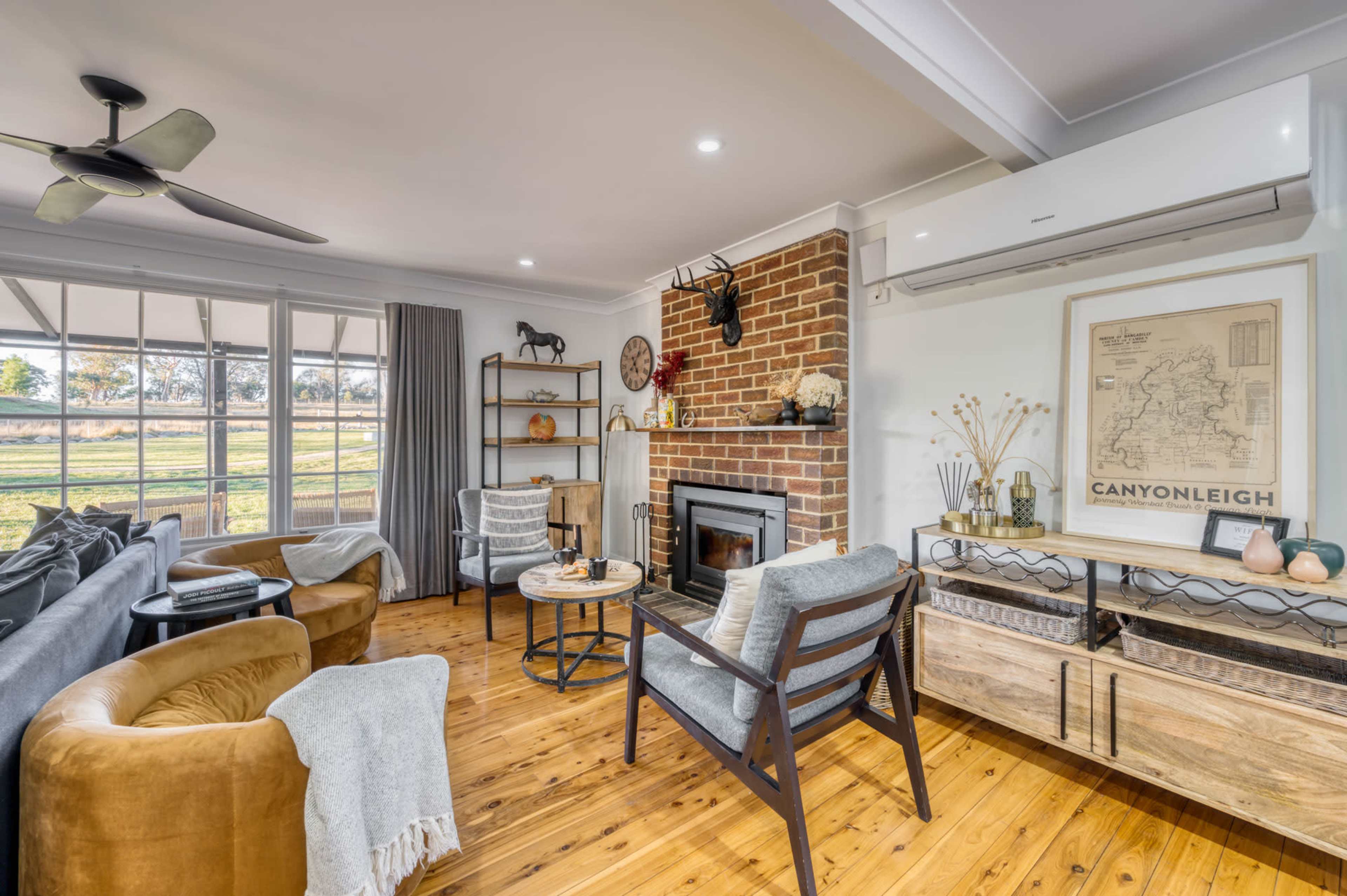 A cozy living room features wooden flooring, a brick fireplace, and a large window overlooking a scenic view, with various seating arrangements and decorative elements.
