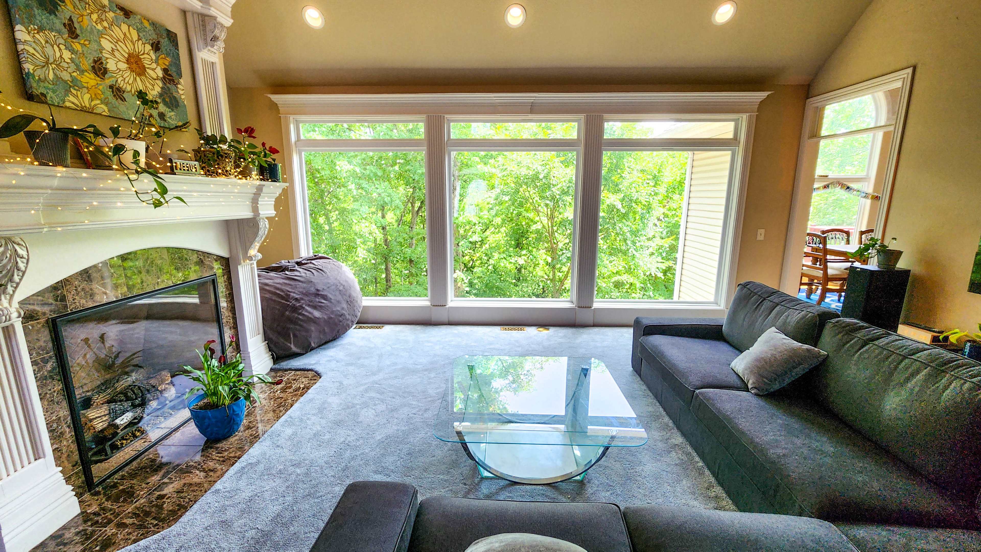 A modern living room features a glass coffee table, gray sofas, a fireplace with a decorative mantle, and large windows overlooking green vegetation.