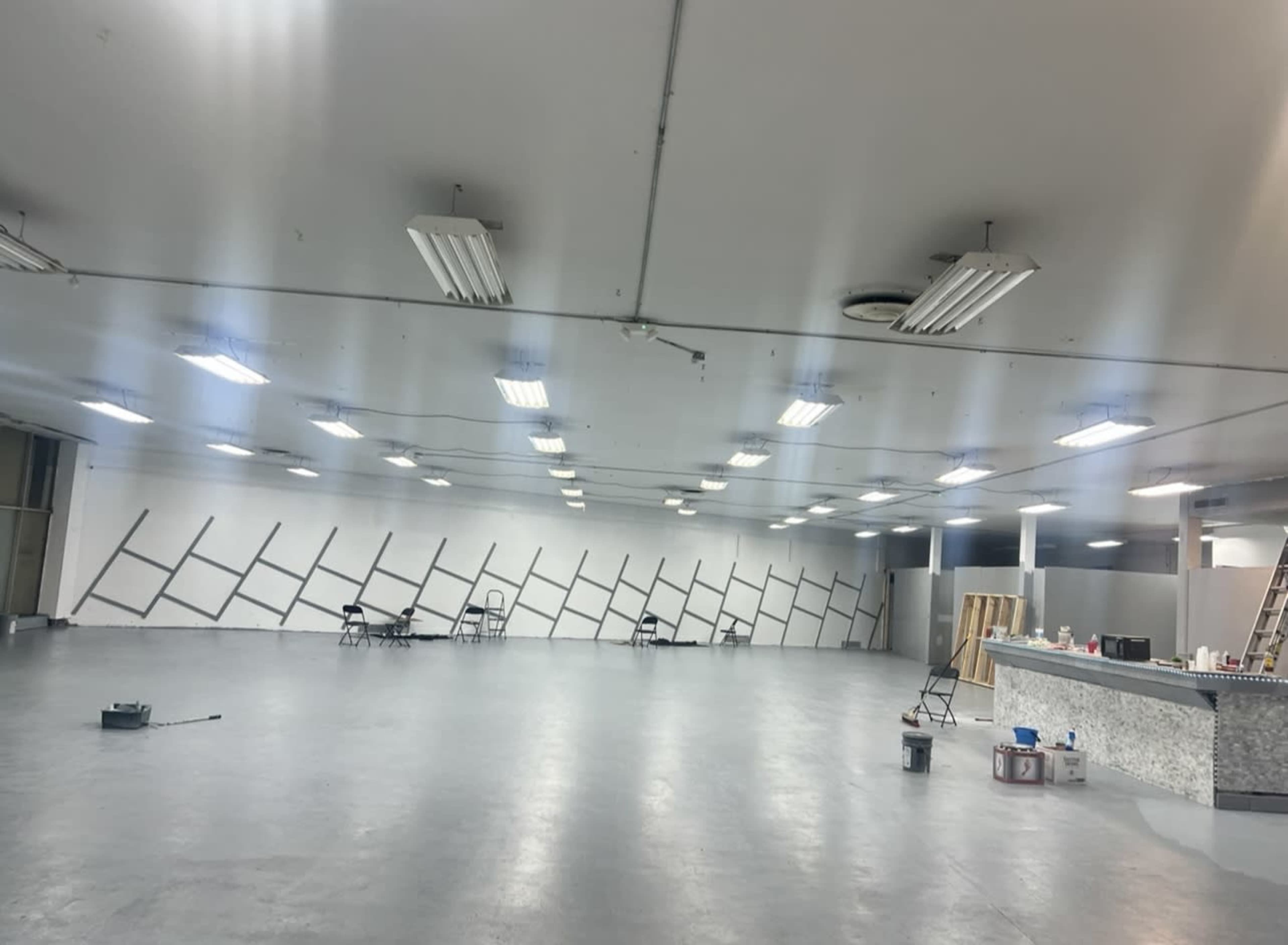 A large, empty interior space with a polished concrete floor, bright fluorescent lights, and a wall featuring diagonal black stripes.