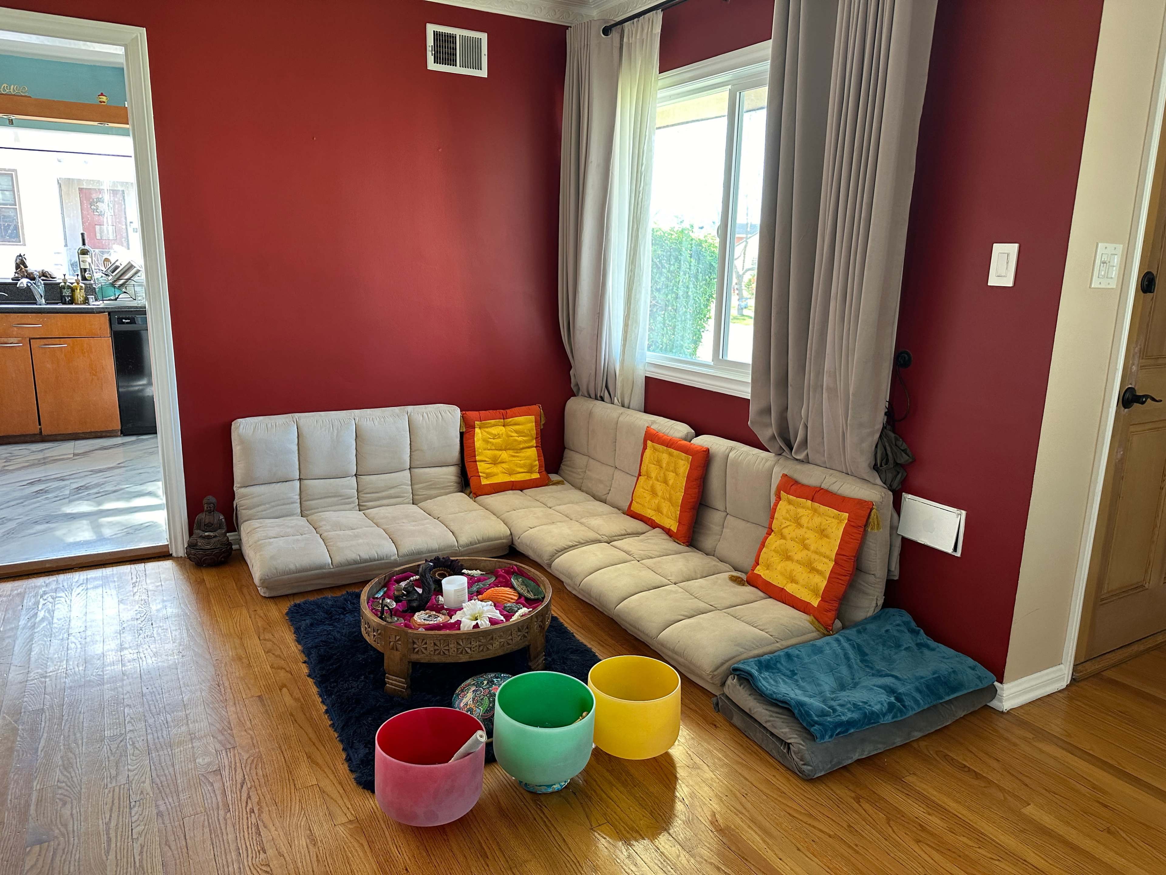 A cozy seating area features a grey modular sofa with orange cushions, colorful bowls, and a decorative tray in a room with red walls and a window.