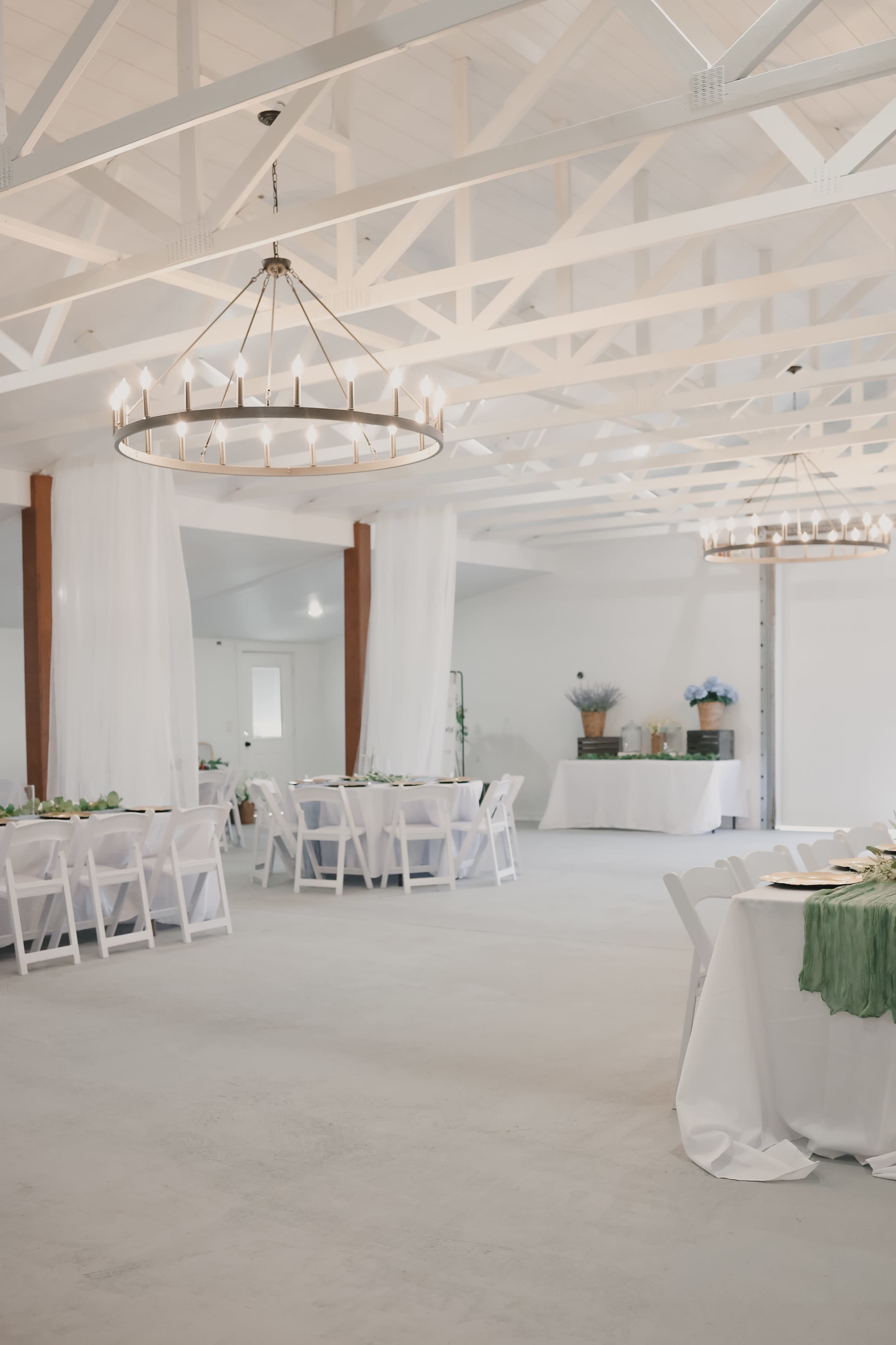 The image shows a spacious event venue with tables set for dining, elegant chandeliers overhead, and light-colored decor.