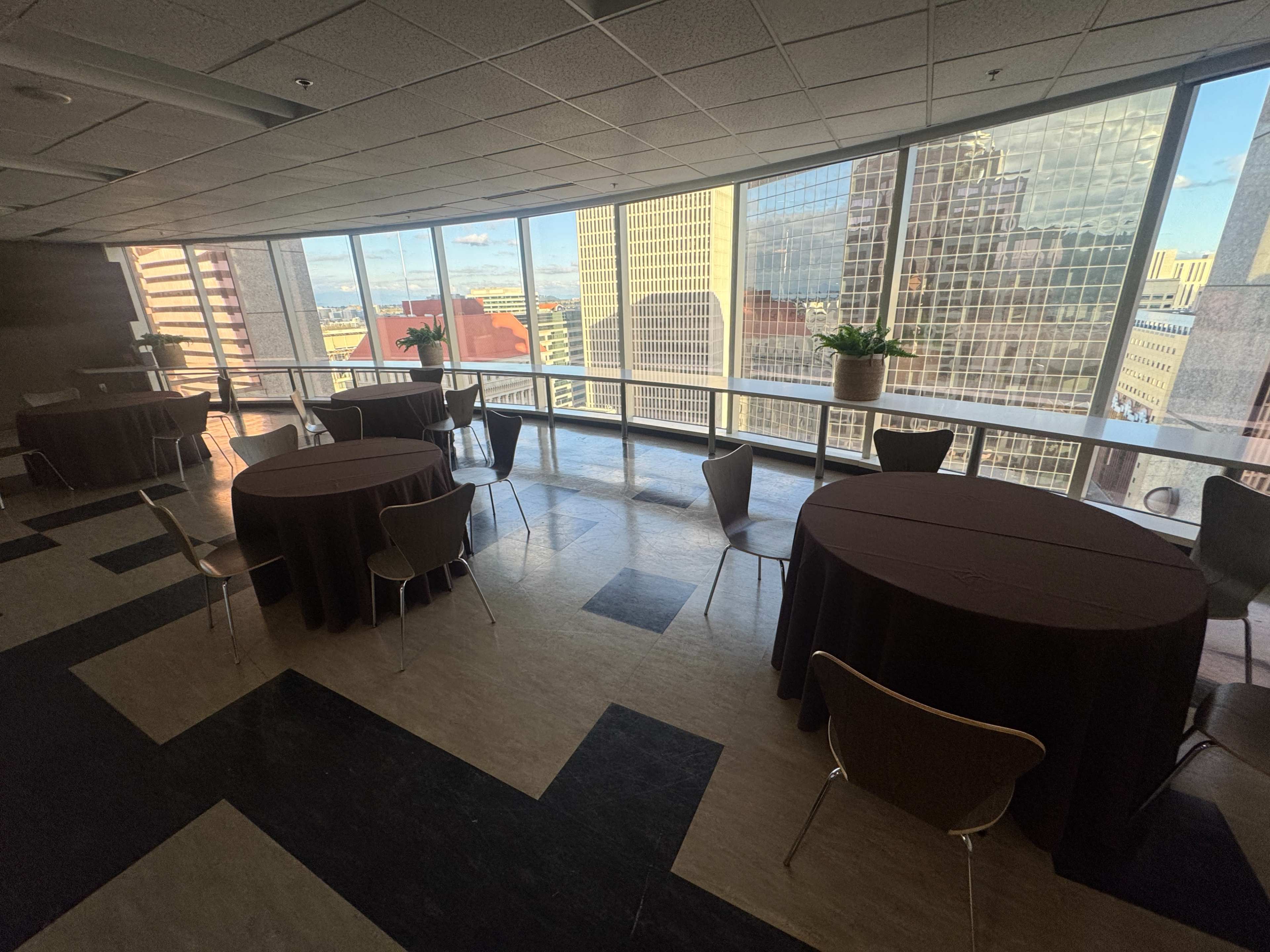 The image shows a spacious indoor area with tables covered in brown cloth and large windows overlooking a cityscape filled with tall buildings.