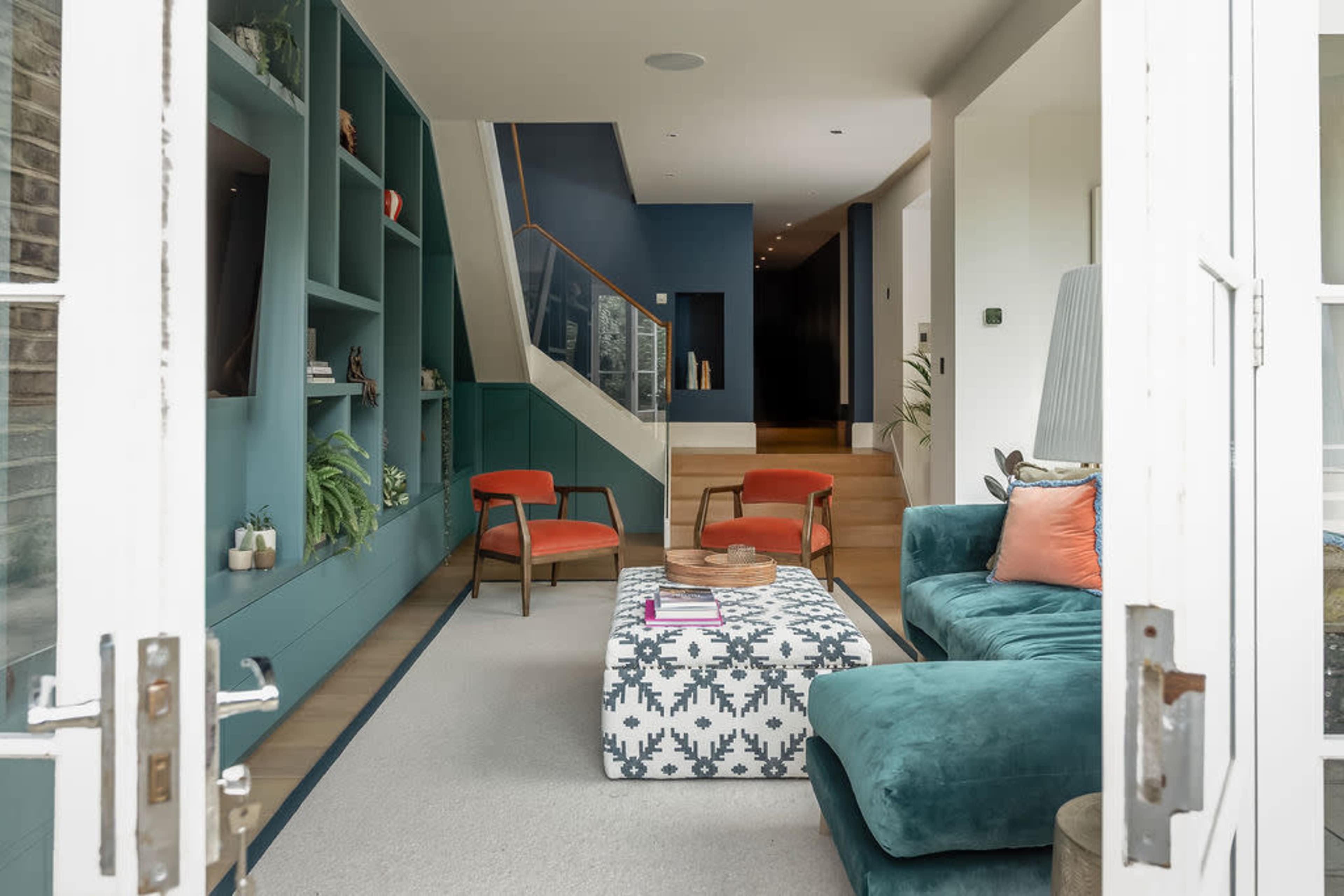 The image shows a modern living room with a teal sofa, patterned coffee table, orange chairs, and a staircase leading to another level.