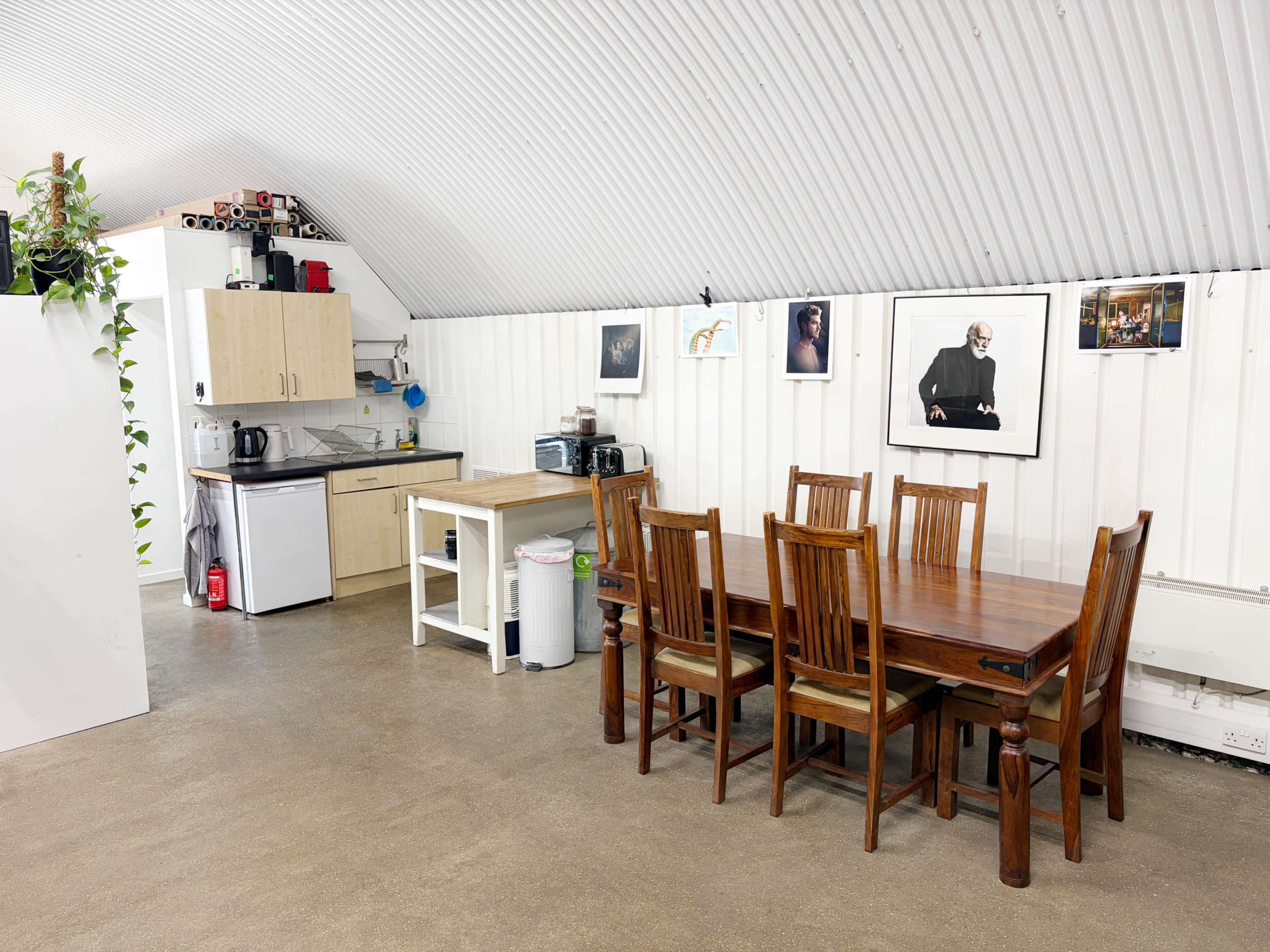 The image shows a dining area with a wooden table and chairs, adjacent to a kitchenette featuring countertops, a sink, and various appliances along a white wall with framed photos.