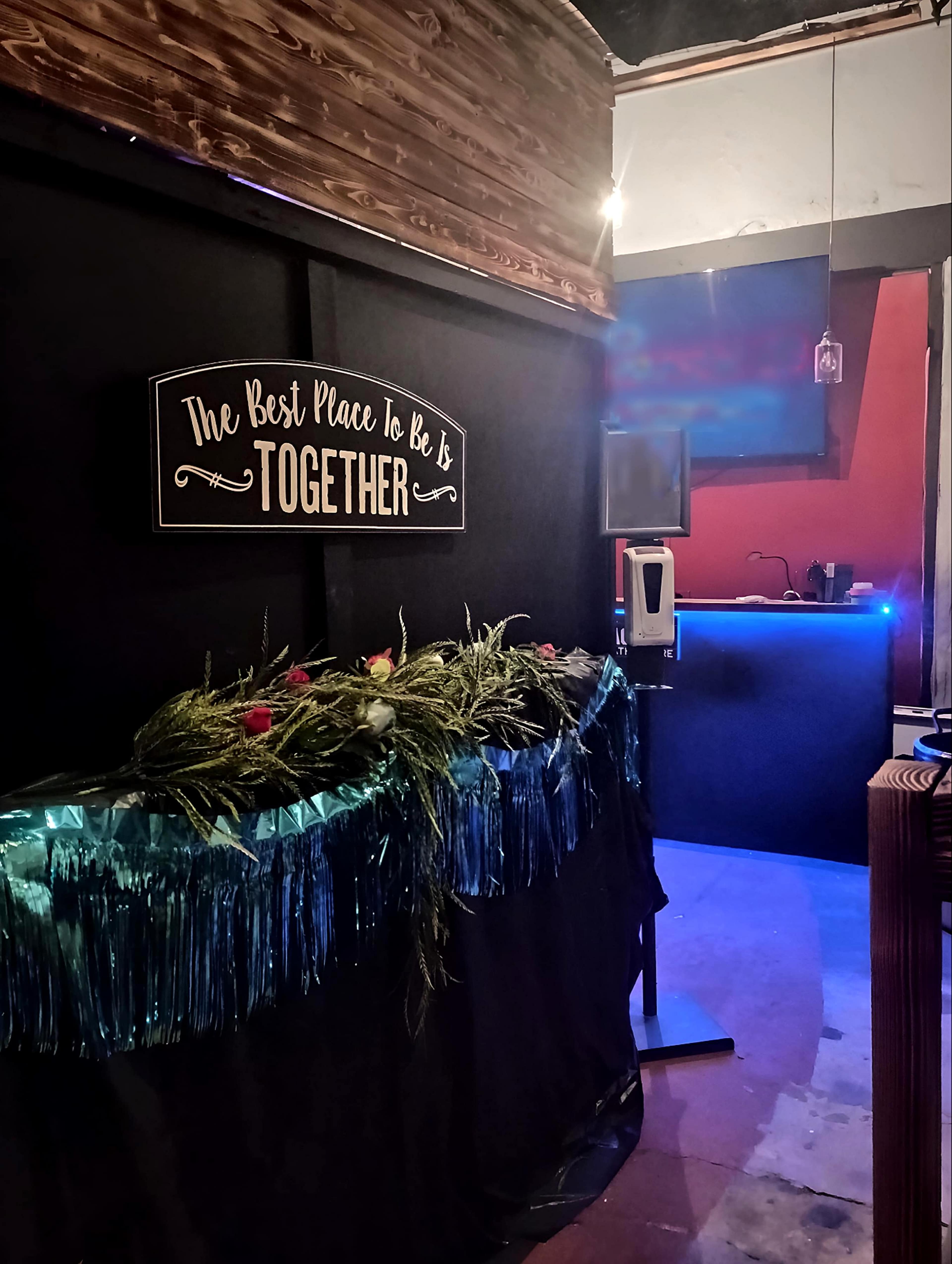 A decorated table with greenery and flowers in front of a sign that reads "The Best Place to Be Is TOGETHER," adjacent to a bar area with blue lighting.