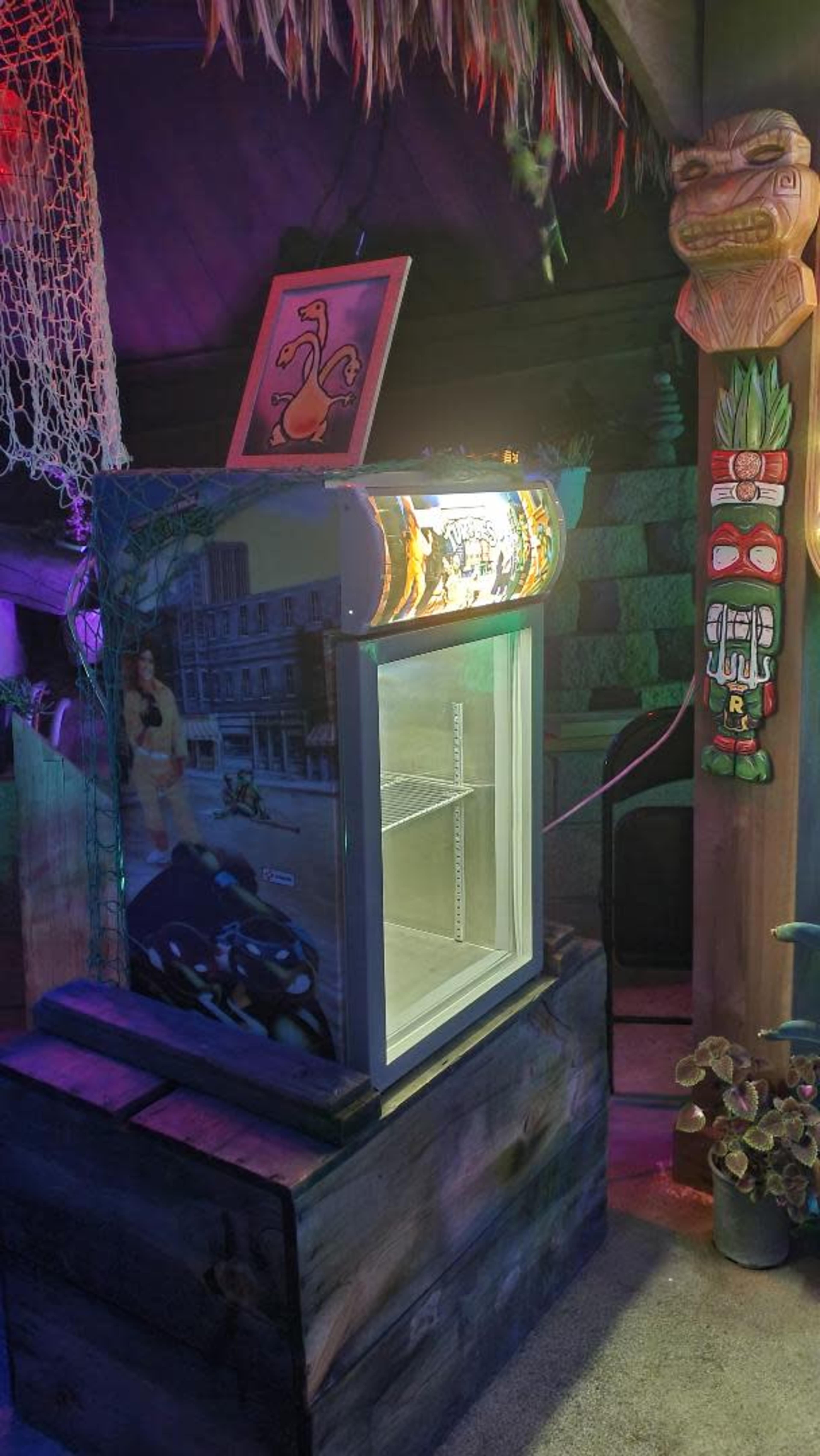 The image shows a colorful mini refrigerator decorated with tropical and cartoon-themed artwork, placed in a dimly lit room adorned with tiki masks and a fishing net.