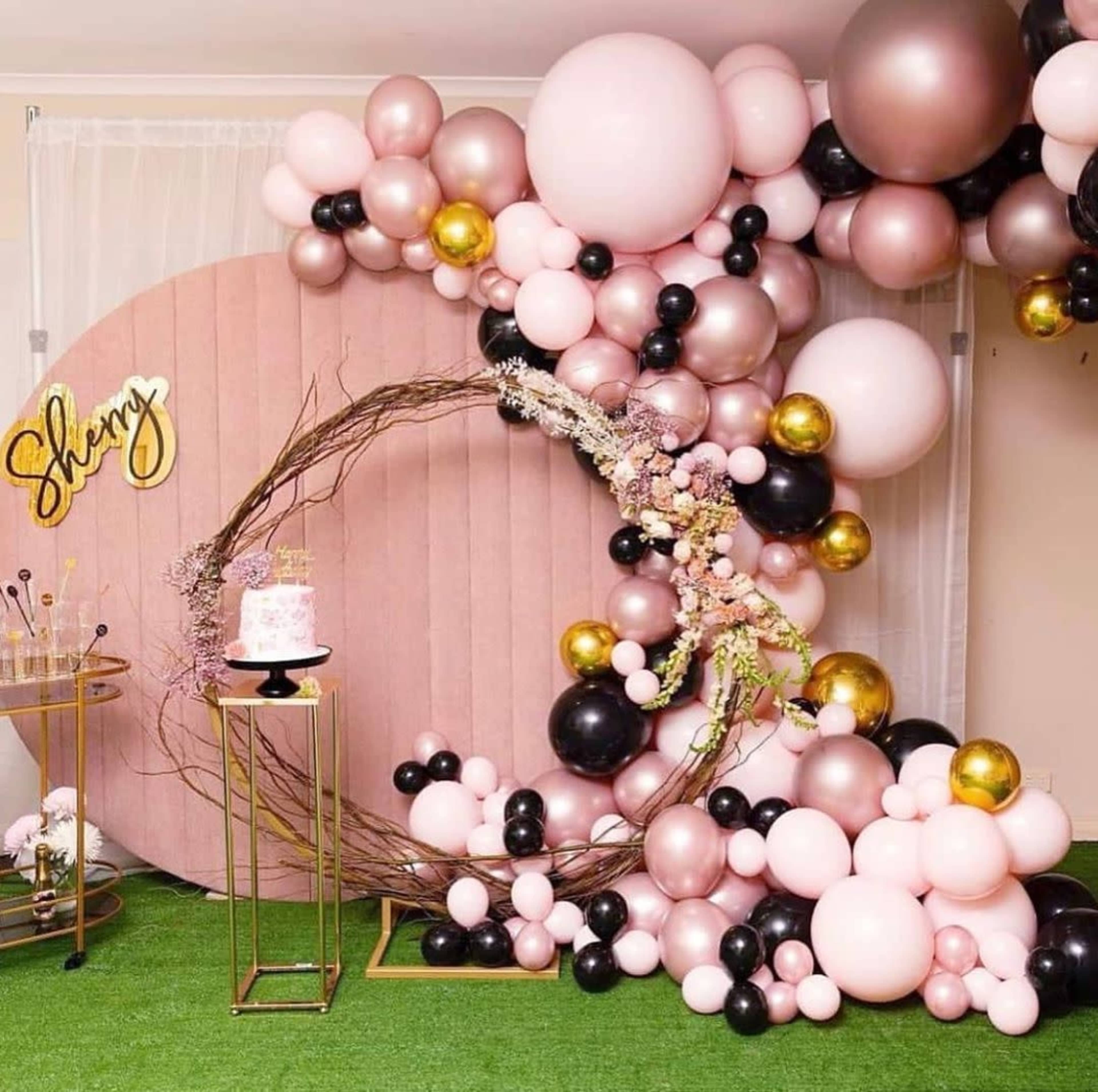 A festive decorations setup featuring a circular balloon arch with pink, black, and gold balloons, a small cake on a gold stand, and a sign that says "Shay" against a backdrop of grass-like flooring.