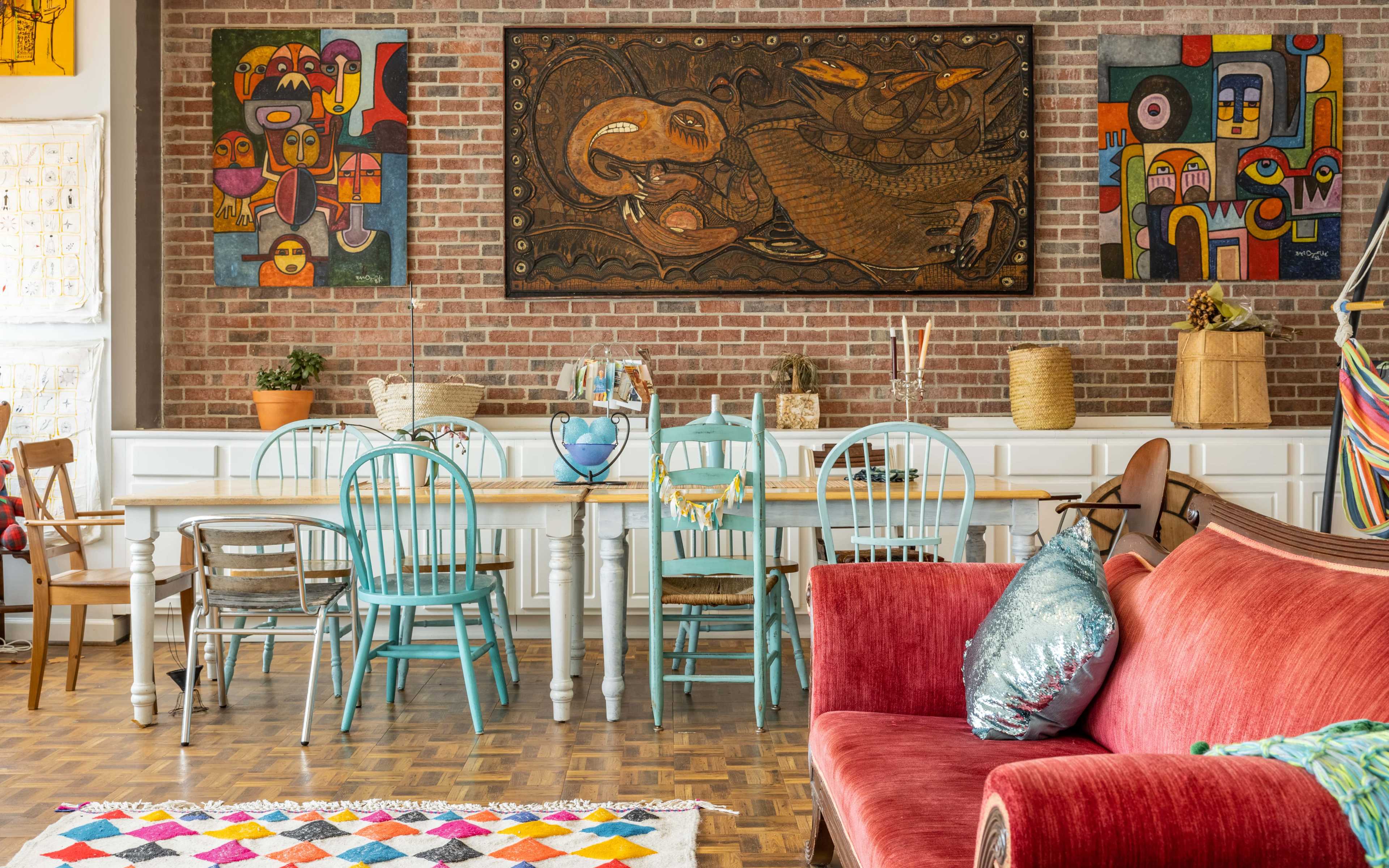 A stylish living room with a red sofa, a wooden dining table surrounded by chairs, and colorful artwork displayed on a brick wall.