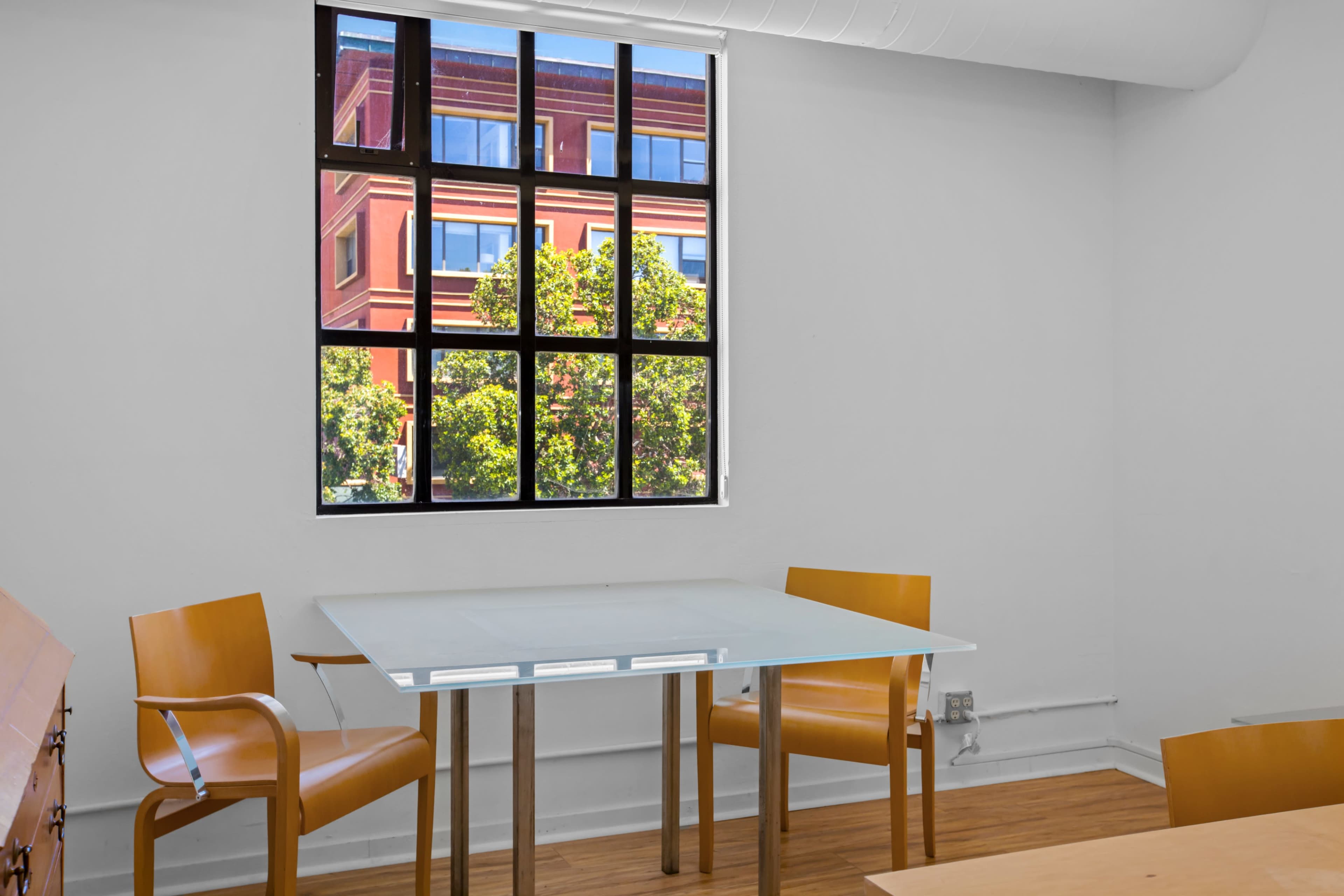 A glass table with two wooden chairs is positioned near a window overlooking a building with trees outside.