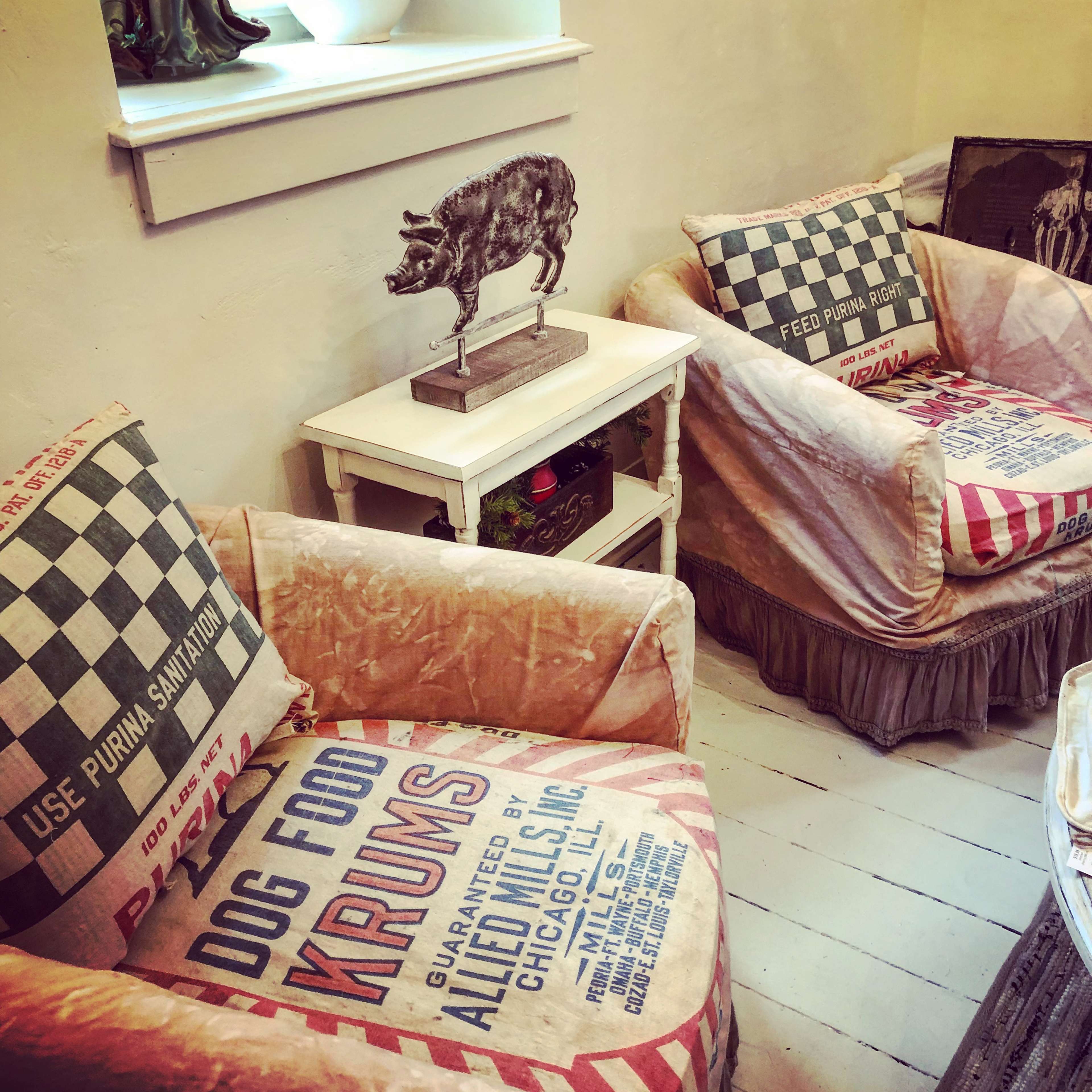 Two vintage chairs with fabric featuring dog food advertisements and decorative pillows sit beside a small table displaying a metal pig statue.