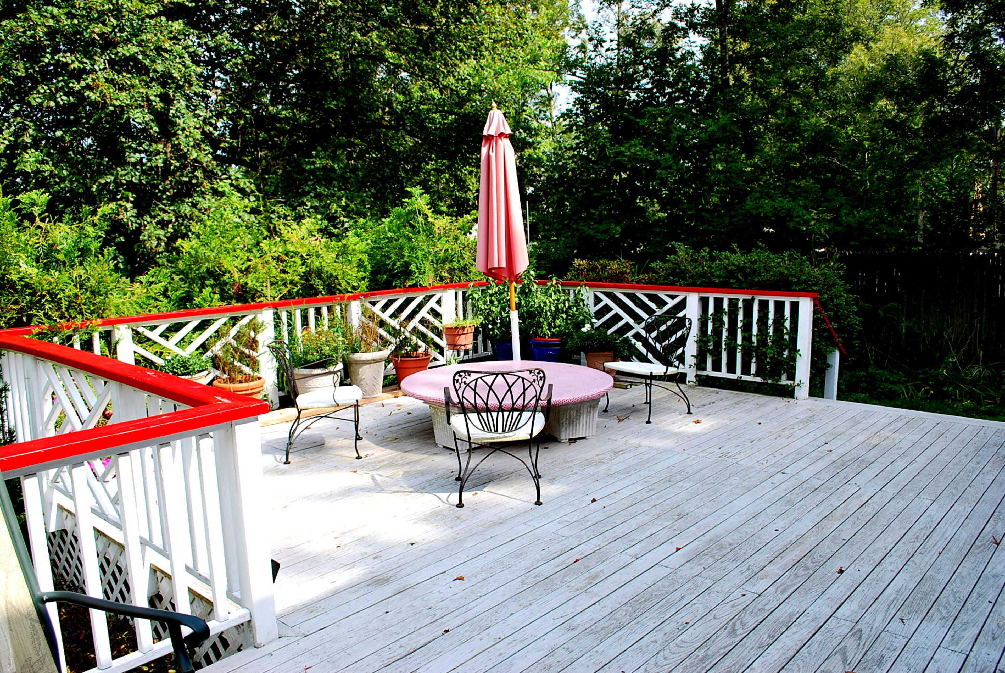 A wooden deck features a round table with a pink tablecloth and six chairs, surrounded by potted plants and a red umbrella.
